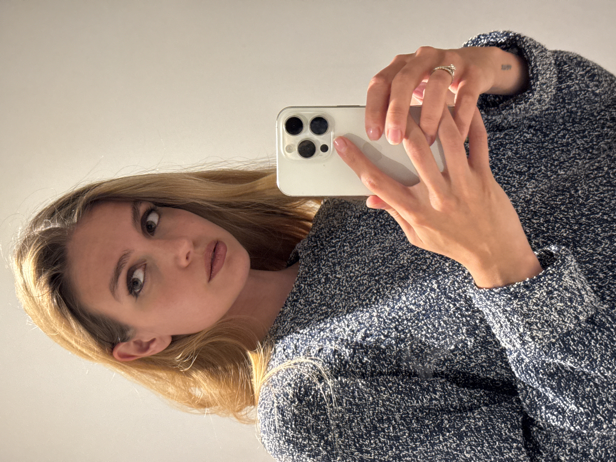 A fair-skinned woman with blonde hair is holding a white cell phone in her right hand, taking a selfie. She wears a navy blue sweater and a diamond ring on her left ring finger. A small tattoo is visible on her left wrist. The background is a solid, light-colored surface.