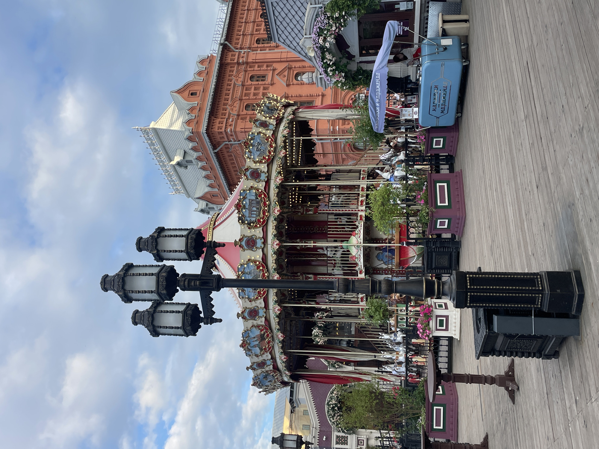 The image shows a carousel in a public space in Moscow, Russia. The carousel is red and white with painted panels depicting scenes from Russian landmarks. It is surrounded by decorative planters and a wooden deck. In the background, a large, ornate orange building, likely a historical structure, stands against a cloudy sky. To the right of the carousel, a blue food cart selling ice cream is visible. A lamppost with three lights stands in the foreground. Several people are present, some near the food cart and others on or around the carousel, but their faces are not clearly discernible. The overall scene suggests a recreational area or park.