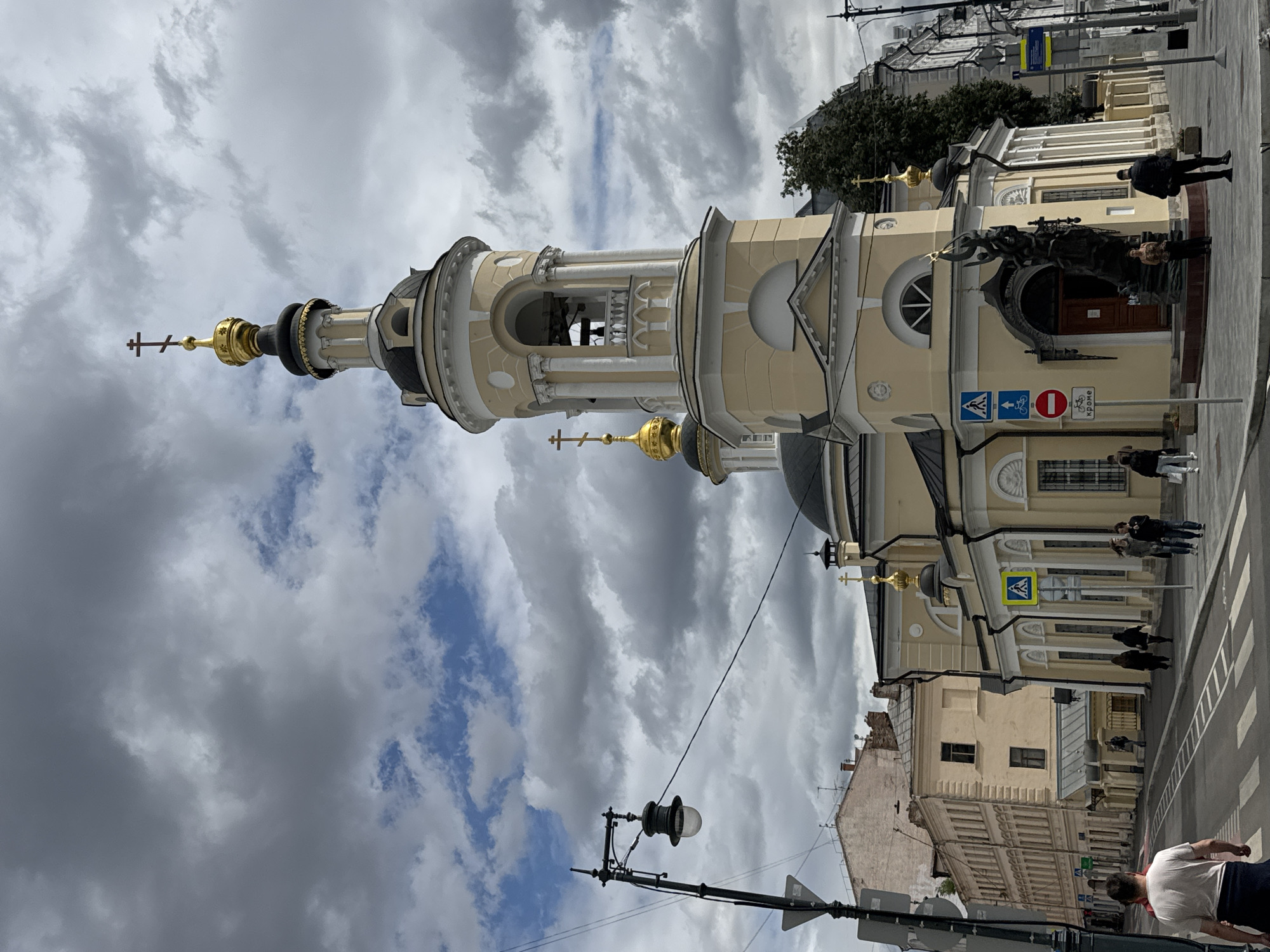 This image depicts the Church of St. Clement Pope of Rome, located in Moscow, Russia. The church features a pale yellow facade with white architectural details and a prominent bell tower topped with a gold dome and a cross. Adjacent to the church, there is a street with a pedestrian crossing, several people walking, and a lamppost. Buildings with varying architectural styles line the street, some with multiple stories and light-colored facades. The sky is dramatic, with a mix of dark, heavy clouds and patches of blue sky, suggesting an overcast day. In front of the church entrance, a large, dark bronze sculpture of figures is visible. Traffic signs, including a pedestrian crossing sign and a prohibition sign, are present along the sidewalk.