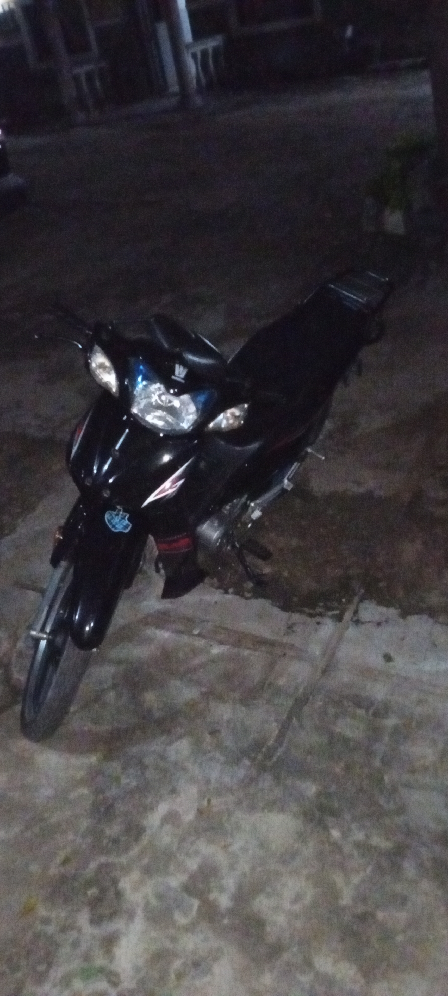 A black motorcycle is parked on a concrete surface outdoors at night. The surrounding area is dimly lit, suggesting a residential or less populated part of the city of Adankolo, Nigeria. The concrete surface has an uneven texture and appears to be a driveway or parking area. Background buildings are visible, but details are obscured by the darkness. A small potted plant stands in the background. The motorcycle is positioned at an angle to the camera, allowing for a view of its front headlight, side panels, and the length of the seat.
