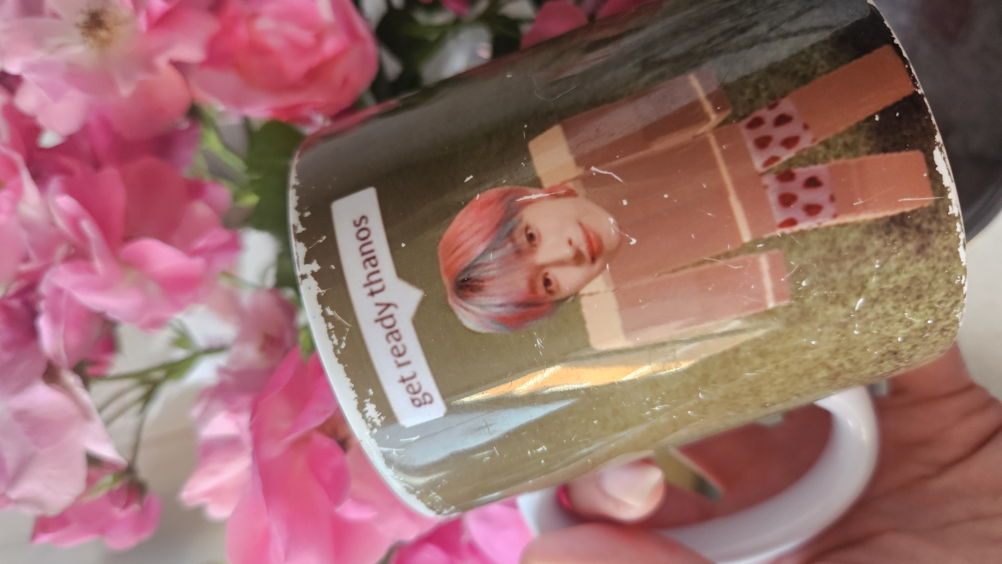 A person's hand holds a mug decorated with a picture of a person with pink and blue hair, and a Roblox-style character wearing shorts with red hearts. A speech bubble above the person's head says "get ready thanos". The mug is positioned in front of a bouquet of pink roses. The background is blurred. The image does not contain any information about the city of Krasnodar, Russia.