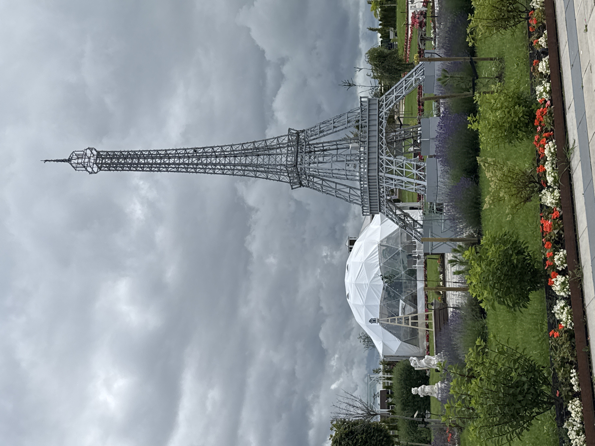 The image captures an outdoor scene featuring a miniature Eiffel Tower and a geodesic dome structure. The Eiffel Tower replica is a metallic, lattice-like structure with intricate detailing, standing prominently in the center-right of the frame. To its left is a large, white geodesic dome, partially covered by a translucent material, with a ladder leaning against it. The foreground is a well-maintained garden with green grass, hedges, and flowerbeds containing red and white flowers. Two white statues of figures, possibly mythological or decorative, are placed in the garden. The background shows more greenery, trees, and a cloudy, overcast sky. The scene appears to be in a park or an amusement area, identified as being located in Bruzdeilynas, Lithuania. There are no people visible in the image.