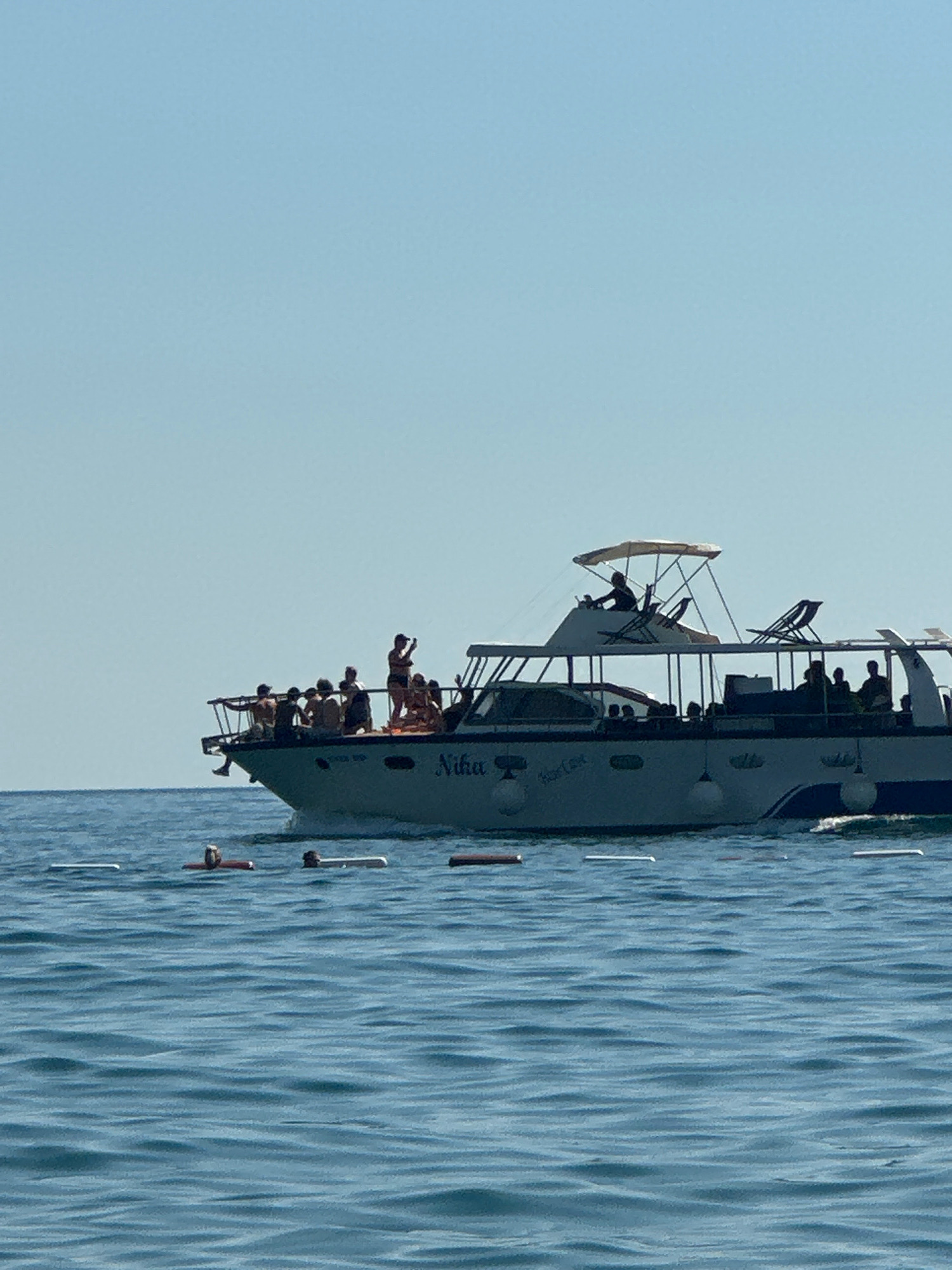 This image depicts a pleasure boat named "Nika" with a group of people on board, sailing in the sea near Boreti, Montenegro. The sky is clear and blue, and the water is a vibrant blue with visible ripples and some floating buoys in the foreground. The boat is a white, multi-level vessel with a covered top deck where a person is seated at the helm, and several chairs are arranged. The main deck is populated with approximately 15 individuals, some standing and some seated, appearing to be enjoying a boat trip. Some passengers are at the railing, looking out at the sea. The boat has the name "Nika" visible on its side, along with other text that appears to read "Star Line." The overall scene suggests a recreational outing on a sunny day.