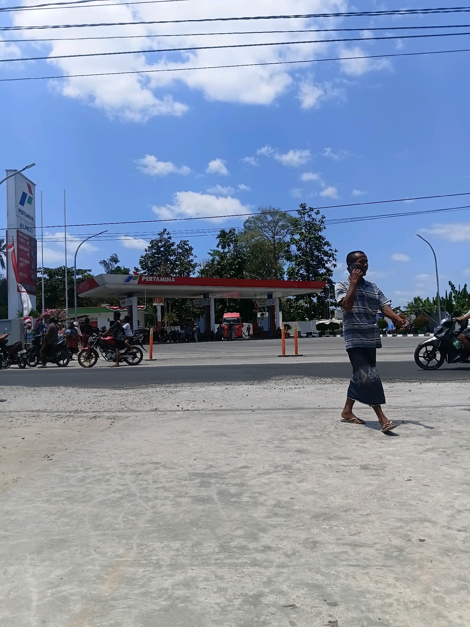 The image depicts a sunny day at a Pertamina gas station in Sungai Kapitan, Indonesia. In the foreground, a man walks while talking on his phone, wearing a striped shirt and a sarong. He is on a paved area, with a street and a gas station visible in the background. The gas station features red and white structures with the Pertamina logo. Several motorcycles are parked and in use at the station, with some people attending to them. The sky is blue with scattered white clouds, and trees are visible in the distance. Power lines are strung across the upper portion of the image.