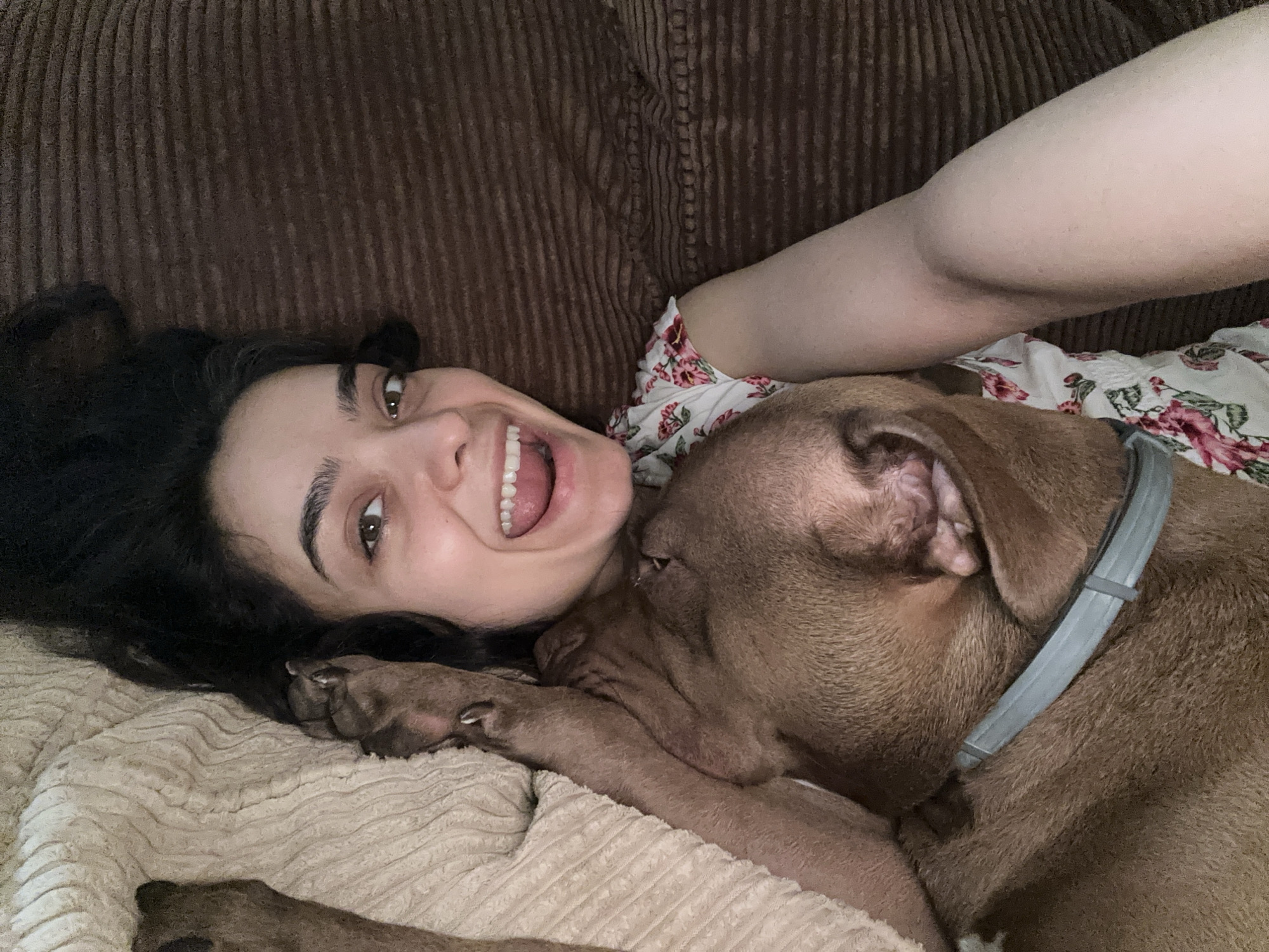 This media file depicts a close-up, indoor selfie of a young woman and a brown dog lying down together on a couch. The woman has dark, wavy hair and is looking up towards the camera with a wide smile, her mouth open and teeth visible. Her eyes are open and her eyebrows are dark. She is wearing a white top with a floral pattern featuring pink and red flowers. The dog, appearing to be a pit bull or a similar breed, is nuzzling against the woman's face. The dog has brown fur and is wearing a grey collar. The background shows the ribbed texture of a brown couch and a textured cream-colored blanket or rug beneath them. The lighting suggests it is daytime, likely indoors, and the mood is intimate and affectionate, capturing a moment of shared comfort between the woman and her pet. There is no visible text in the image.