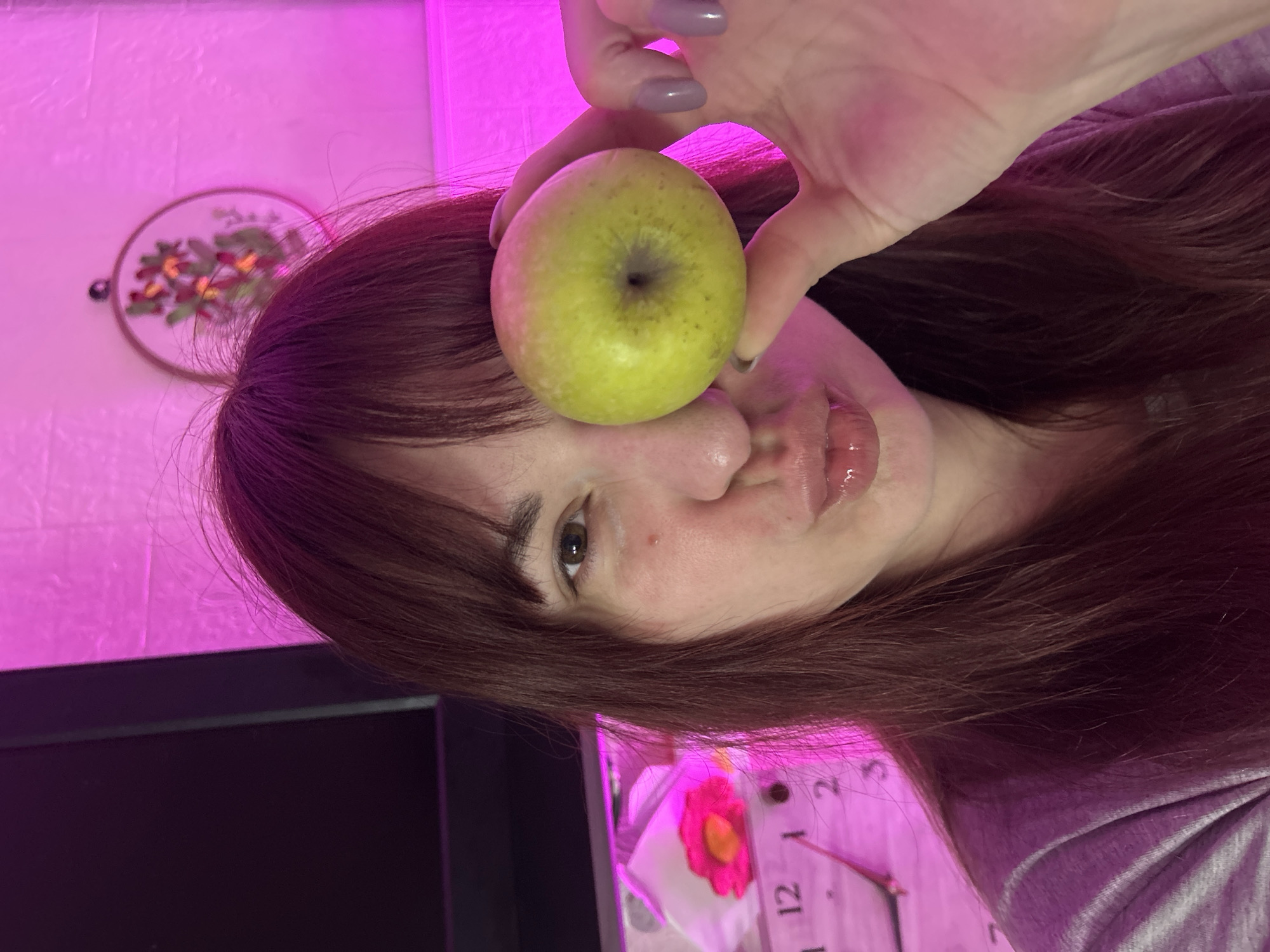 The image shows a close-up portrait of a person in an indoor setting, presented as if rotated 90 degrees counter-clockwise from an upright view. A person with long, reddish-brown hair is prominent, holding a light green apple with their left hand. Their hand, featuring light purple nail polish, positions the apple over one of their eyes, while their visible brown eye looks towards the viewer. The person's full lips are glossy and slightly pouted. They are wearing a light-colored top.
The background is dominated by a strong pink or magenta artificial light, illuminating a textured wall. On the wall, a blurred circular embroidered piece of art with floral patterns in red, orange, and green can be seen. In the lower right section of the image, a portion of a clock face is visible, displaying the numbers '1' and '2', along with a pink flower decoration. A dark, rectangular object, possibly a monitor or TV screen, is situated in the lower left corner.
The scene captures a playful or whimsical moment, likely a casual portrait, within a home interior. The artificial pink lighting creates a distinct and modern atmosphere. The provided context indicates the location is Фоки, Russia.