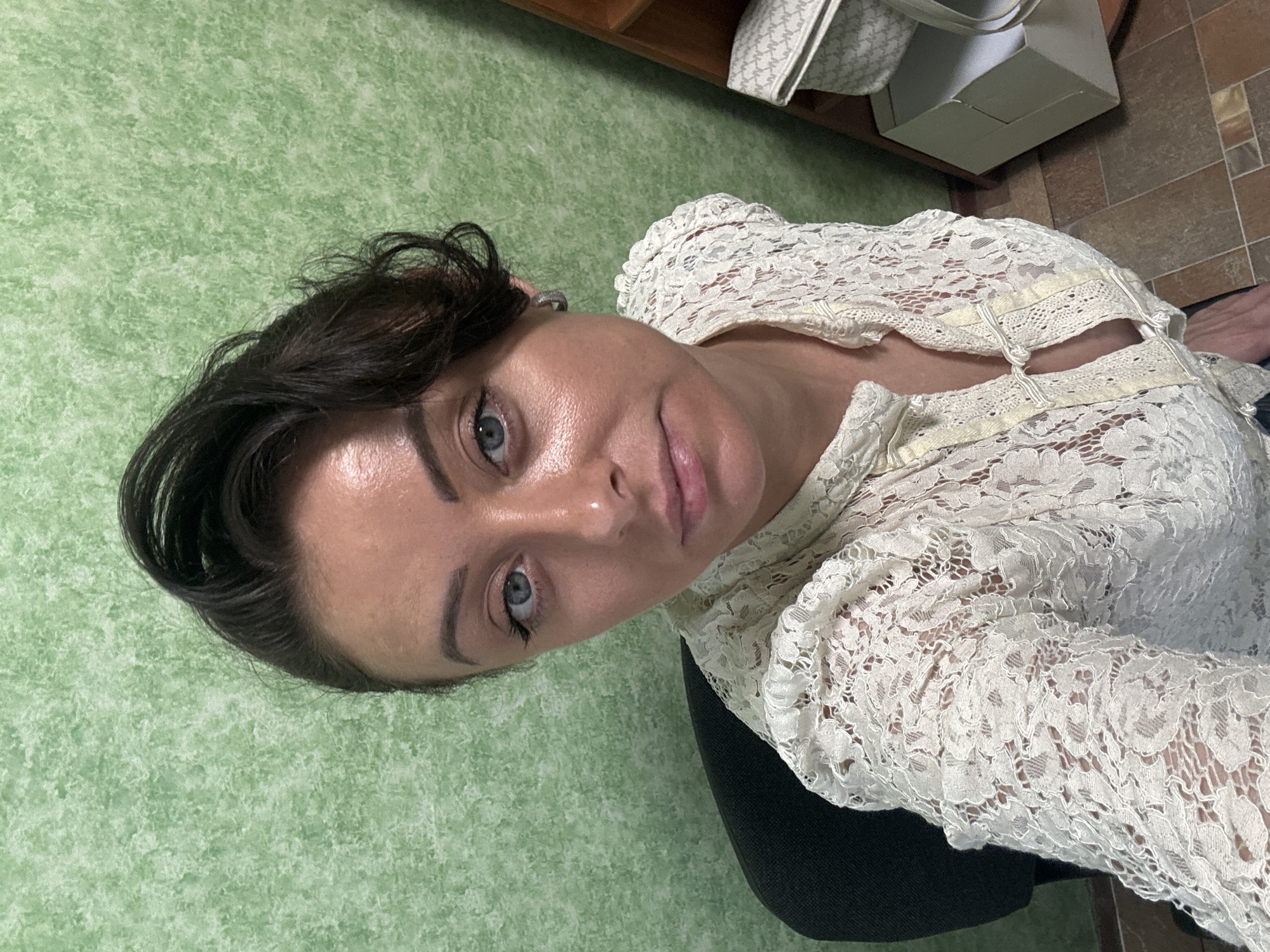 An overhead, medium shot captures a woman with dark hair and blue eyes, seated in a black office chair, looking directly at the camera. She is wearing a cream-colored lace blouse with a high neckline and a decorative knot closure. The background features a textured green wall. To the right, a wooden shelf unit holds a white tote bag with a monogram pattern and a light-colored box. The floor is tiled with a mosaic of brown, tan, and beige squares. The lighting is bright, with the subject's face illuminated from the front.
