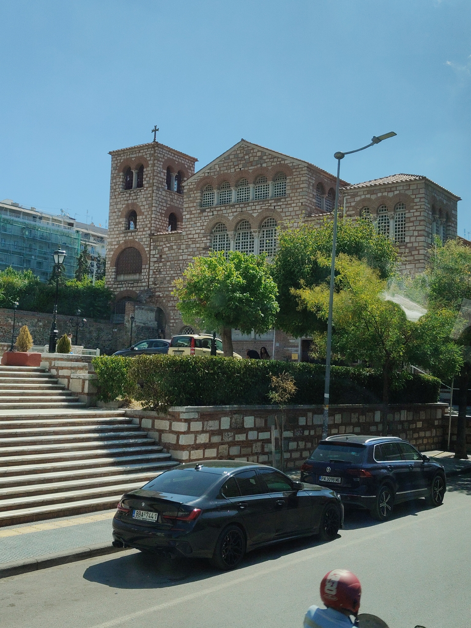 The image depicts a daytime scene in Thessaloniki, Greece, likely near the Hagia Sophia Church. In the foreground, a black BMW sedan is parked on a street. Behind it, a dark blue Volkswagen SUV is also parked. A person wearing a red helmet is partially visible in the lower right corner, suggesting they are on a motorcycle or scooter.
The background features a large, historic stone building with a bell tower, characteristic of Byzantine architecture, which appears to be Hagia Sophia. The building is adorned with arched windows and intricate stonework. To the left of the church, a wide staircase with white steps leads up to a manicured garden area with hedges and small trees. Further in the background, modern buildings, some under construction with scaffolding, are visible. The sky is clear and blue, indicating fair weather. The overall environment suggests an urban setting with a mix of historical and modern elements.