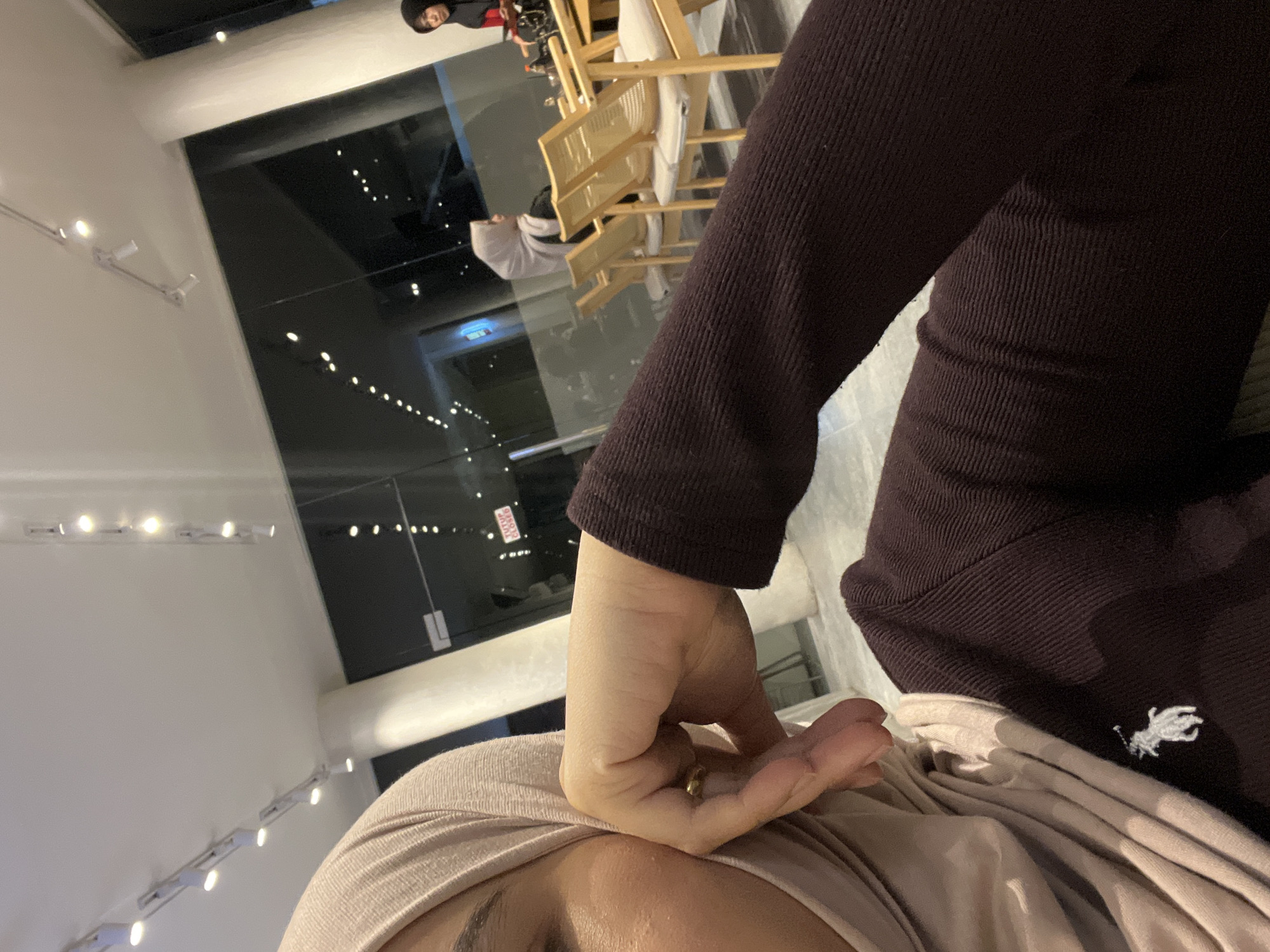 The image shows a close-up of a person wearing a light beige hijab and a dark maroon long-sleeved shirt. The person's right hand is resting on their chin, with a gold ring visible on one finger. A small white embroidered logo, possibly of a horse and rider, is present on the shirt. In the background, through a large glass window, other individuals are seated at tables in what appears to be a restaurant or cafe. One person in the background is wearing a light grey hijab and another is wearing a dark hijab with a red collar. Chairs are visible, and the interior lighting suggests it is evening or night. A sign that reads "TUTUP CLOSED" is visible on the window. The overall environment appears to be a modern indoor establishment. The location is specified as Lhokseumawe, Indonesia.