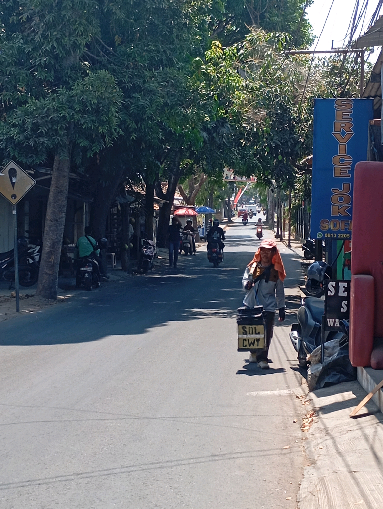 The image shows a street scene in Cimahi, Indonesia, on a bright, sunny day. In the foreground, a person wearing a grey jacket, an orange cloth draped over their head and shoulders, and glasses is walking towards the viewer. They are carrying a black box with yellow lettering that reads "SOL CWY." The person is walking on the right side of the road.
To the right of the person, a large blue sign with white lettering is visible, displaying "SERVICE JOK." Below that, a phone number "0813 2205" is printed. Adjacent to the sign is a red object, possibly a piece of furniture or a structure.
The street is paved and appears to be a two-way road with some parked motorcycles on both sides. Trees with dense green foliage line the street, providing shade in some areas. Several other individuals are seen on motorcycles further down the road, suggesting active traffic. A yellow diamond-shaped sign with a black border and a black exclamation mark inside is visible on the left side of the road, indicating a warning. Shops or small businesses are present along the left side of the street, with some of their structures and awnings visible. The overall atmosphere is that of a typical urban street in Indonesia, with a mix of pedestrian and vehicle activity.