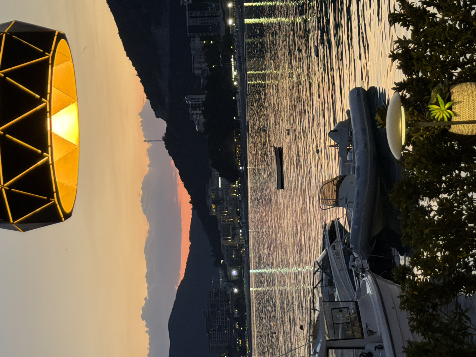 View of Rafailovići, Montenegro at dusk from an elevated, waterfront position. Boats are docked in the water, with the city lights reflecting on the water's surface. A geometric lamp is positioned on the left, and a tree obscures part of the scene on the right. The landscape includes a mountain range.