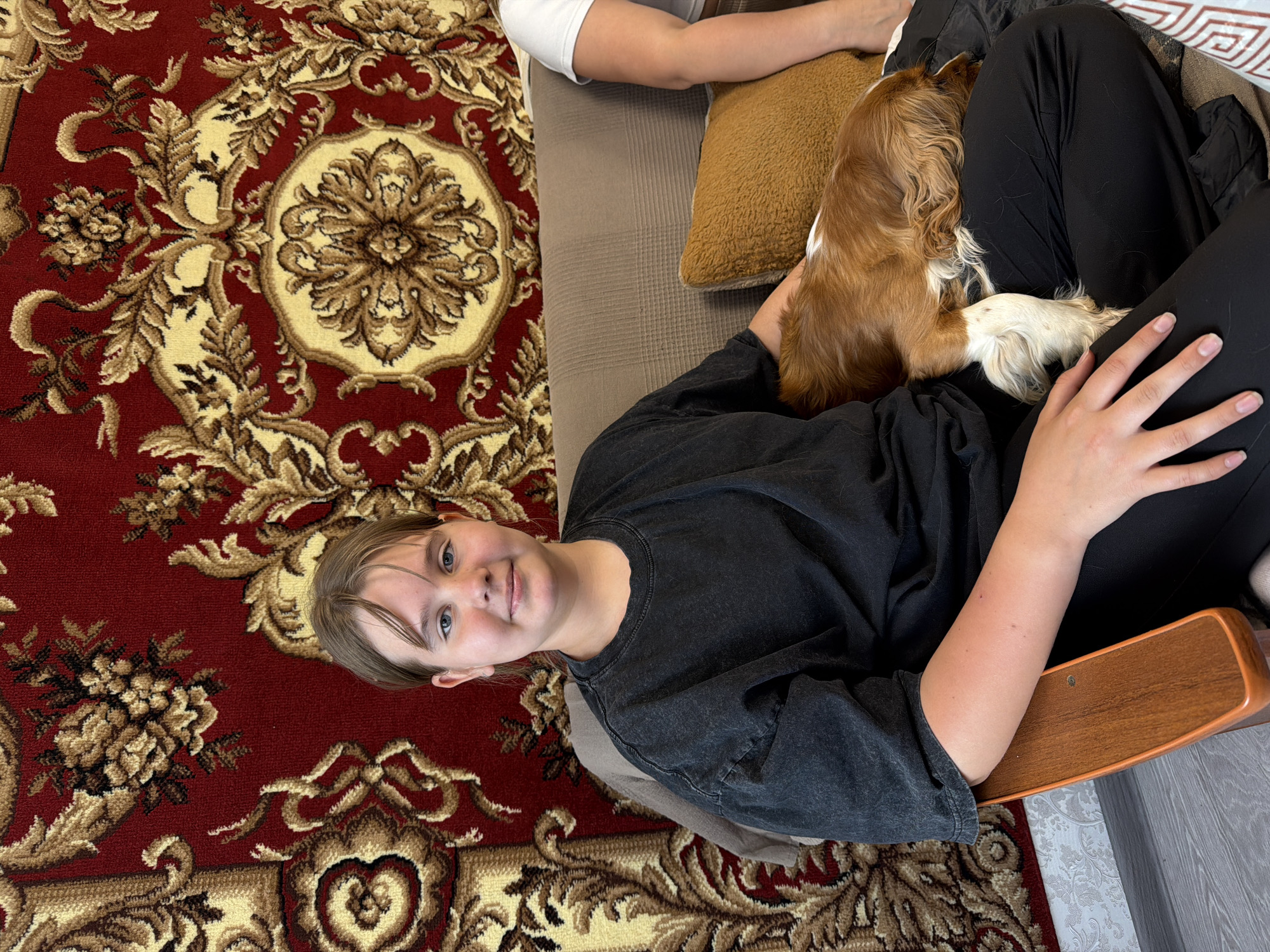 A photograph taken indoors depicts a young person reclining on a sofa or sectional, with a Cavalier King Charles Spaniel resting on their lap. The young person is wearing a dark, possibly charcoal-colored, long-sleeved shirt and dark pants. The dog has primarily brown and white fur. The seating area appears to be shared by another person, evidenced by a portion of a white shirt and arm visible in the upper portion of the frame.
The sofa is positioned atop a patterned rug with a predominantly red background interspersed with gold floral or botanical designs. A brown cushion is visible adjacent to the dog. A wooden chair arm is situated at the bottom-right corner of the frame. The location is Бронница, Russia.
