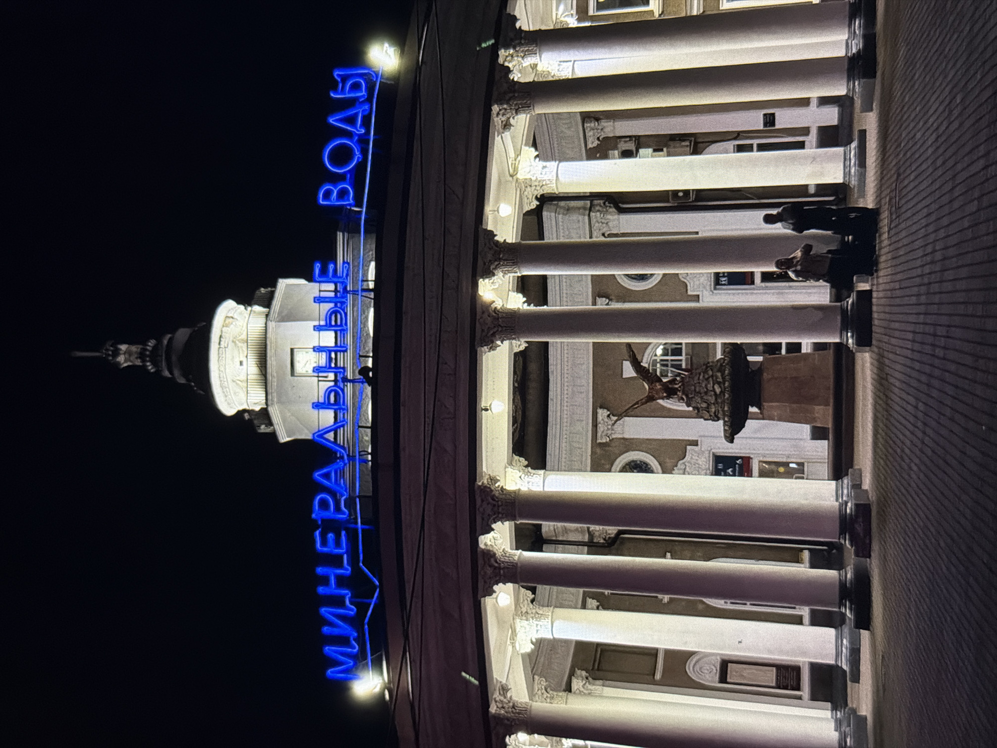 The image shows the entrance of the Mineralnye Vody railway station at night. The station is located in the city of Mineralnye Vody, Stavropol Krai, Russia, not Krasnodar. A large neon sign spelling "МИНЕРАЛЬНЫЕ ВОДЫ" (Mineral Waters) in blue is illuminated above the entrance. The station features a grand facade with a circular colonnade of white pillars. In front of the building, there is a statue of an eagle on a pedestal. Two figures, silhouetted against the light, stand in the foreground on a brick-paved area. The lighting is dramatic, casting long shadows and highlighting the architectural details of the station.