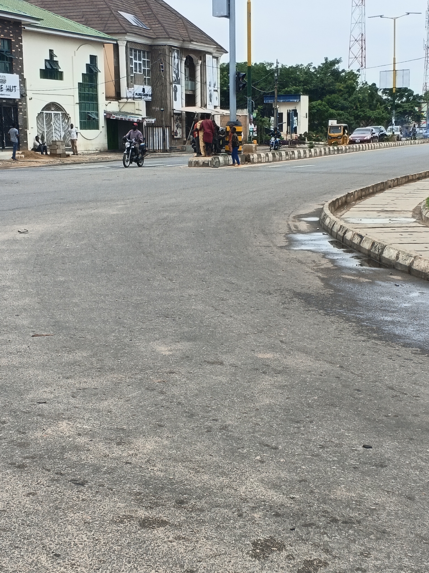 The image captures a street scene in Kabala, Nigeria. The foreground is dominated by a grey asphalt road, showing wear and spots. To the right, a curb and sidewalk edge the street.
In the middle distance, pedestrians cluster at an intersection with traffic signals, while a motorcyclist rides along the road. Further down the street, a yellow tricycle (Keke Napep) and several cars are visible.
Buildings line the left side of the street, including one with green trim and another with multiple windows and signage. In the background, trees and utility poles are visible. The sky is overcast.