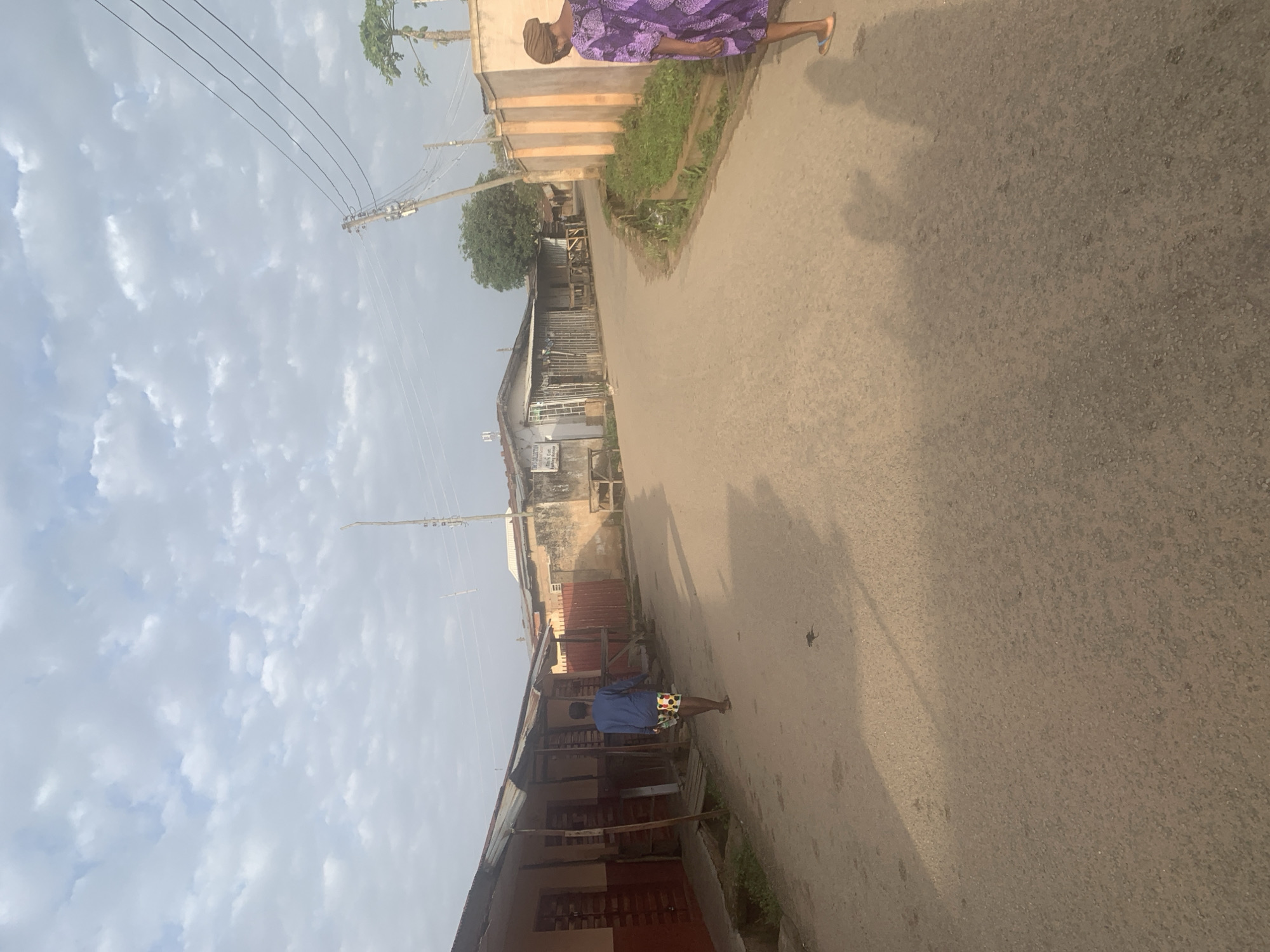 This is a street scene in Jos, Nigeria, depicting a dusty, paved road lined with shops and residential buildings. The sky is partly cloudy, suggesting it may be morning or late afternoon.
On the left side of the road, there are several shops with corrugated metal roofs. One shop has a reddish-brown facade with wooden doors and windows. A person wearing a blue long-sleeved shirt and colorful shorts is walking away from the camera down the middle of the road, casting a shadow.
On the right side of the road, there is a cream-colored wall, likely a boundary for a property, with vertical decorative elements. A woman wearing a vibrant purple patterned dress and a headscarf is standing on the right side of the road, facing away from the camera and slightly towards the road. She is wearing light-colored sandals. Further down the road, more buildings are visible, including structures with corrugated roofs and some concrete buildings. There are power lines and poles present, indicating infrastructure. Green vegetation is visible along the side of the road in some areas. The overall impression is a modest urban environment.