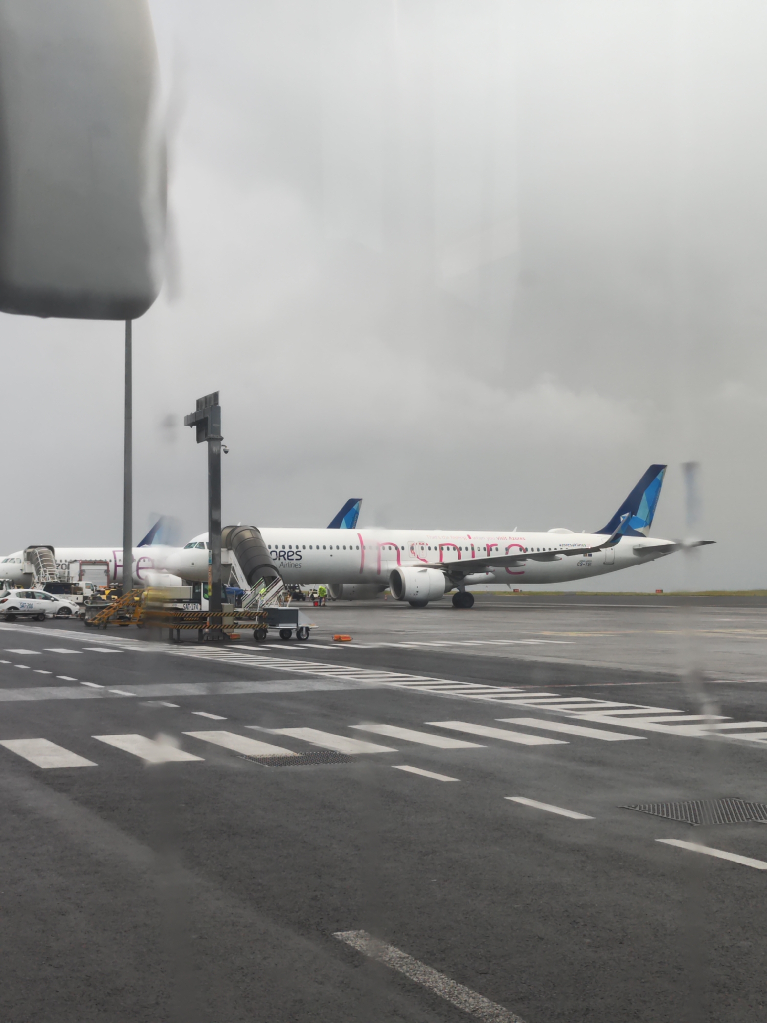 The image captures an overcast daytime scene at an airport tarmac in Ponta Delgada, Portugal. Two passenger airplanes are visible, flanked by ground support equipment.
The prominent aircraft, a white jetliner, features a distinctive livery with pink and purple text, partially reading "Inspire...". It clearly displays "ORES Airlines" and "Visit Azores" on its fuselage, with the registration "CS-TS" near the tail. The tail fin is blue with a lighter blue and white geometric pattern. The right engine of this aircraft shows motion blur, suggesting it might be running or experiencing heat haze.
Behind it, a second white airplane, partially obscured, also shows "ORES Airlines" and "201" on its side. Both aircraft have mobile stairs or jet bridges attached to their doors, indicating active ground operations such as boarding or deboarding. Various yellow and orange ground support vehicles, including baggage carts and a white car labeled "SAT-200," are present on the tarmac around the planes. A black pole structure in the foreground is marked "SAT-174."
The sky is a uniform, heavy gray, reflecting an overcast or rainy day, and the tarmac appears wet. The photograph was taken through a window, with a blurred, reflective object obscuring part of the upper left foreground. The dark asphalt tarmac is marked with clear white lines and pedestrian crossings. The scene depicts typical airport activity under gloomy weather conditions.