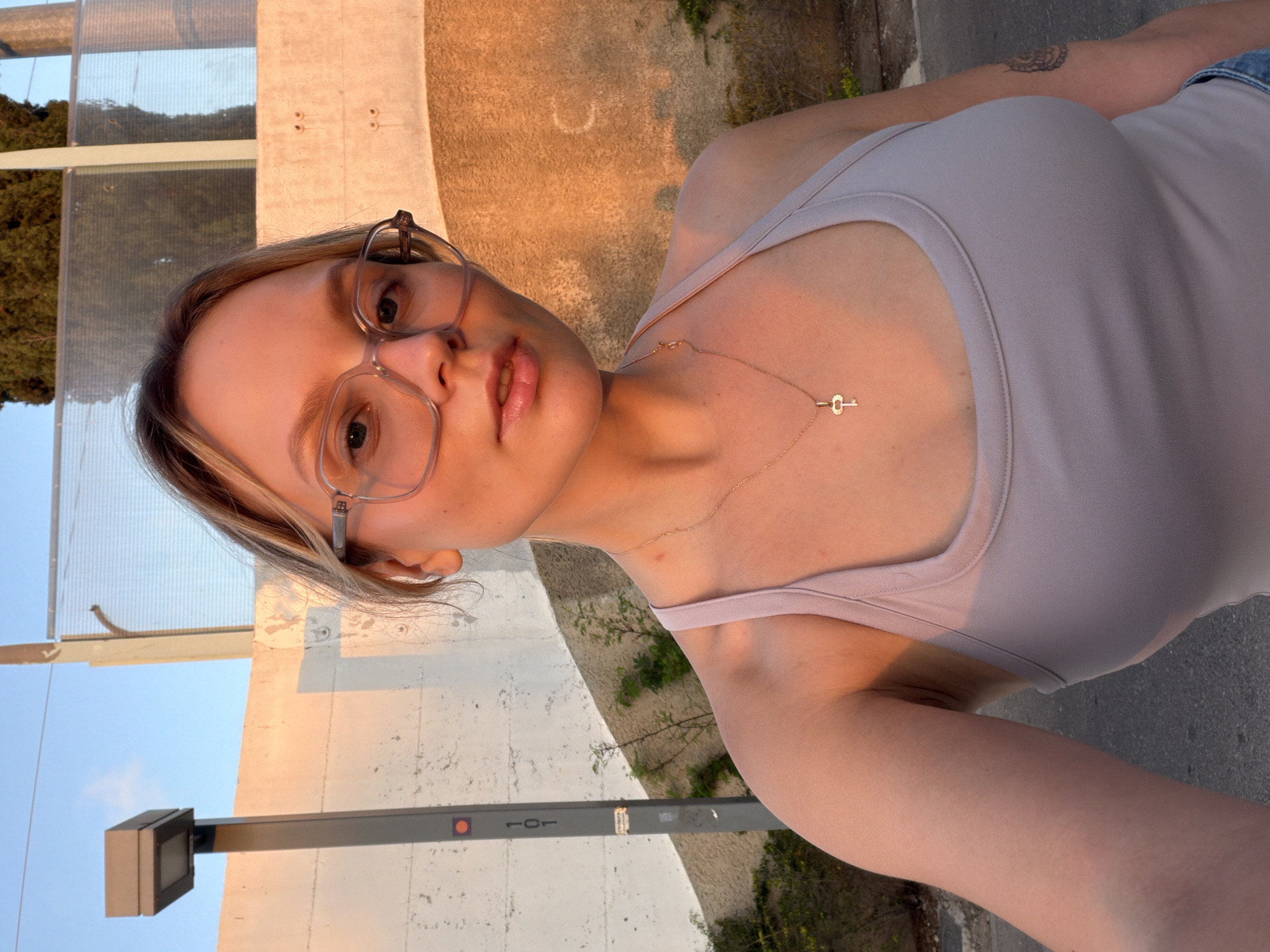 A young woman wearing glasses and a light purple tank top is taking a selfie. She has a key-shaped necklace on. The background shows a concrete wall and a street lamp, with trees and a fence visible further back. The setting sun casts a warm glow on the scene. The image does not contain any specific indicators of it being located in Tel-Aviv, Israel.
