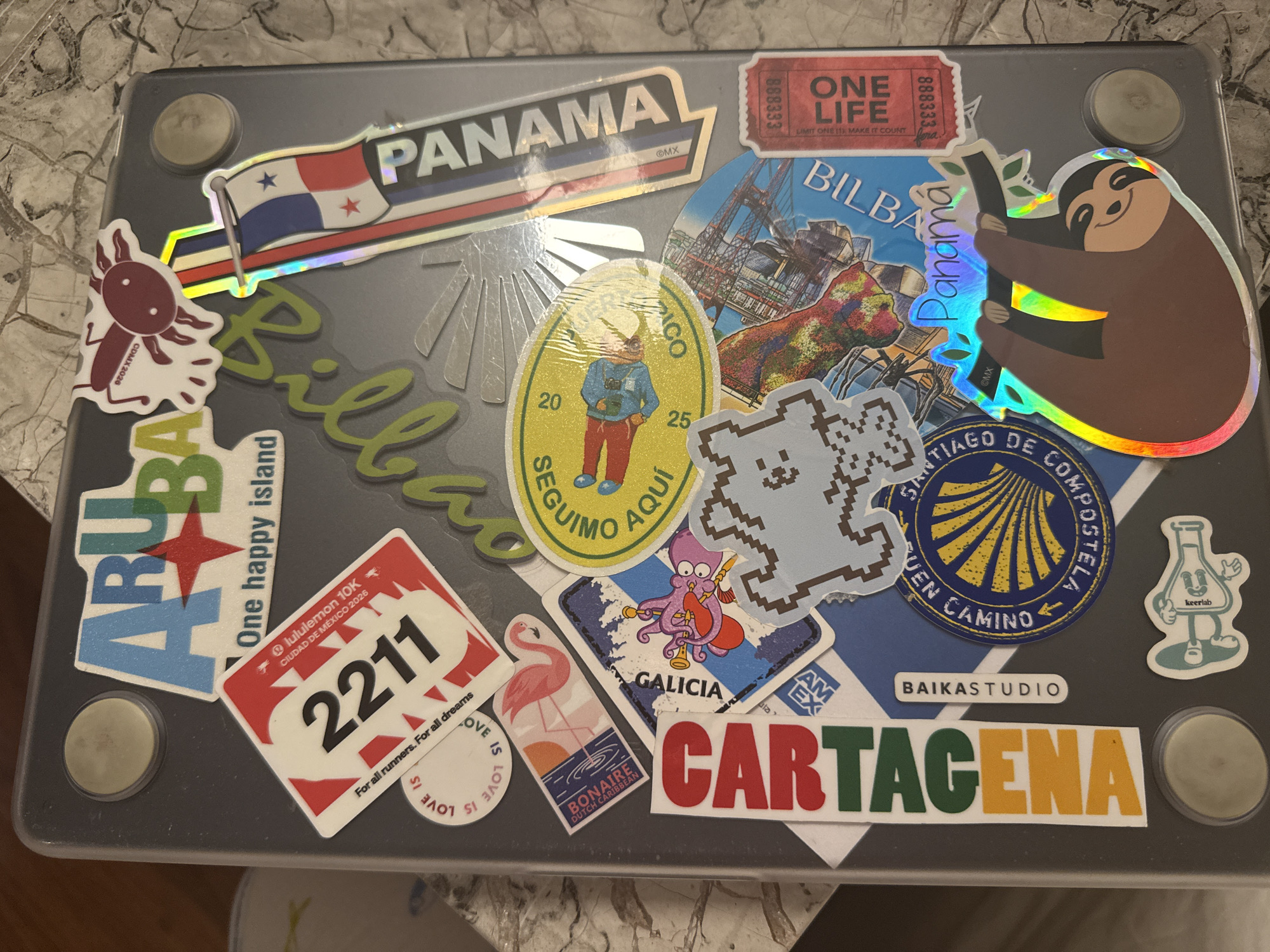 The image shows a gray laptop covered in stickers. The stickers feature various destinations and themes, including: "Panama", "Bilbao", "Aruba", "Ciudad de Mexico 2026", "Bonaire", "Cartagena", "Santiago de Compostela", and "Galicia". There's a "One Life" ticket sticker, a sticker of a sloth hanging from a tree, and other stickers of animals and logos. There are four circular rubber feet on the laptop. The surface beneath the laptop is a mottled stone-like material. There is no identifiable location within San Bernardino Tlaxcalancingo, Mexico.