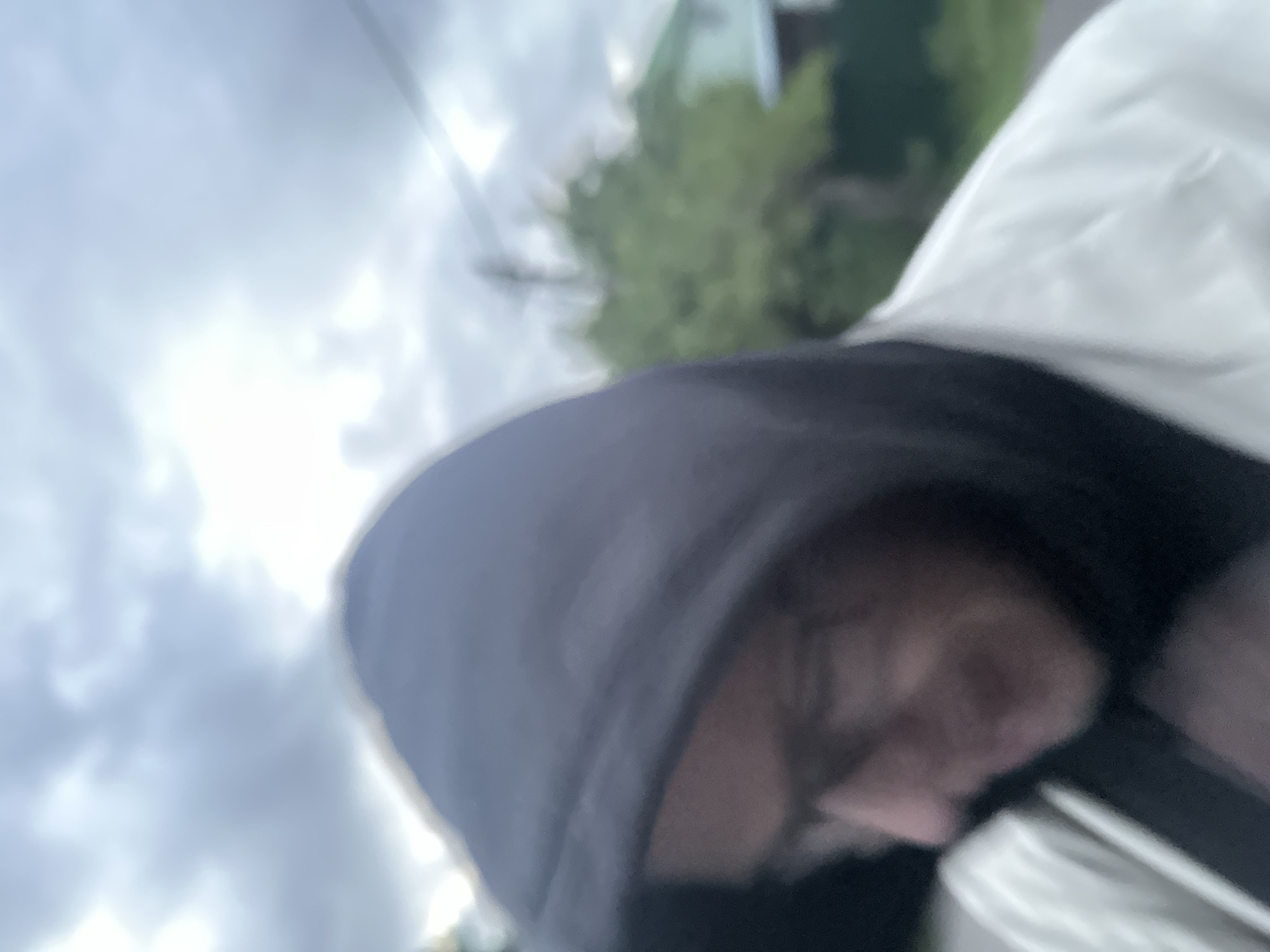 The media file is a close-up, blurry photograph of a person wearing a dark hooded garment. The camera angle is low and tilted, suggesting it may have been taken from the person's perspective or from a low vantage point. The background is out of focus but shows hints of greenery, possibly trees, and a building, which could be a house or other structure, with a light blue hue. The sky appears cloudy and overcast, with some bright patches indicating daylight, possibly late afternoon or early morning given the diffused light.
The person's face is partially visible under the hood, with their head tilted down. Their features are indistinct due to the blurriness of the image and the shadow cast by the hood. The overall impression is casual, possibly candid, with no specific activity clearly depicted beyond the person being outdoors. The presence of what appear to be trees and a building suggests an outdoor, possibly residential or semi-rural setting. The weather seems to be cloudy but not actively raining. No text is visible in the image. The location context provided is "Востец Снт, Russia."