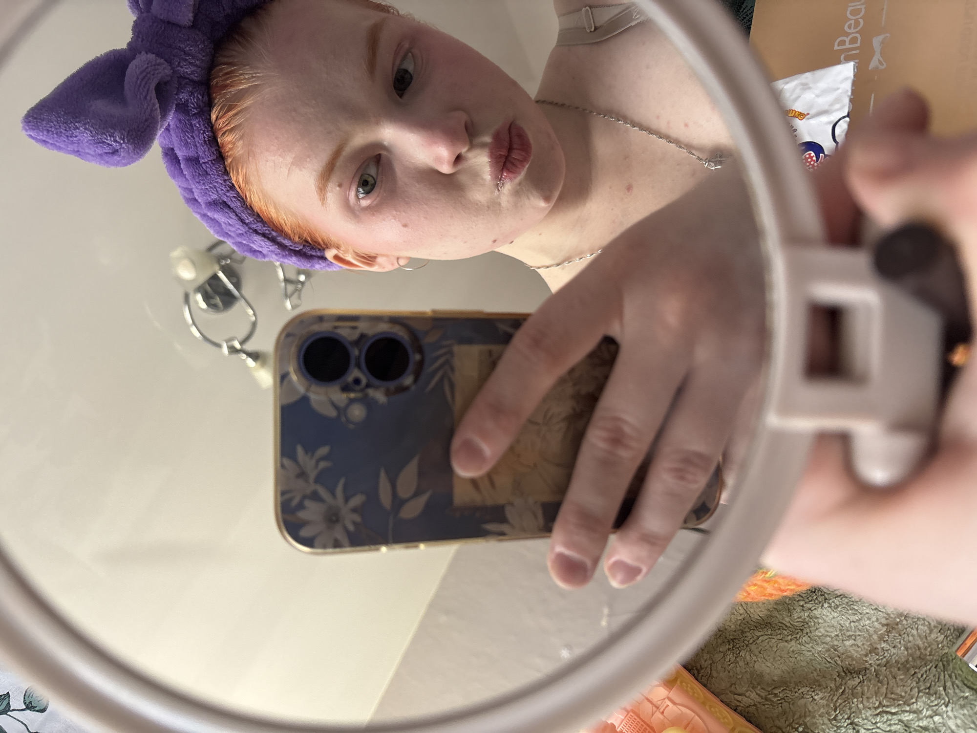 A person with red hair and a purple headband is taking a selfie in a mirror. They are holding a cell phone with a floral case. A silver necklace is visible on their neck. The background includes a white wall and other objects, partially obscured by the mirror. The location is unknown.