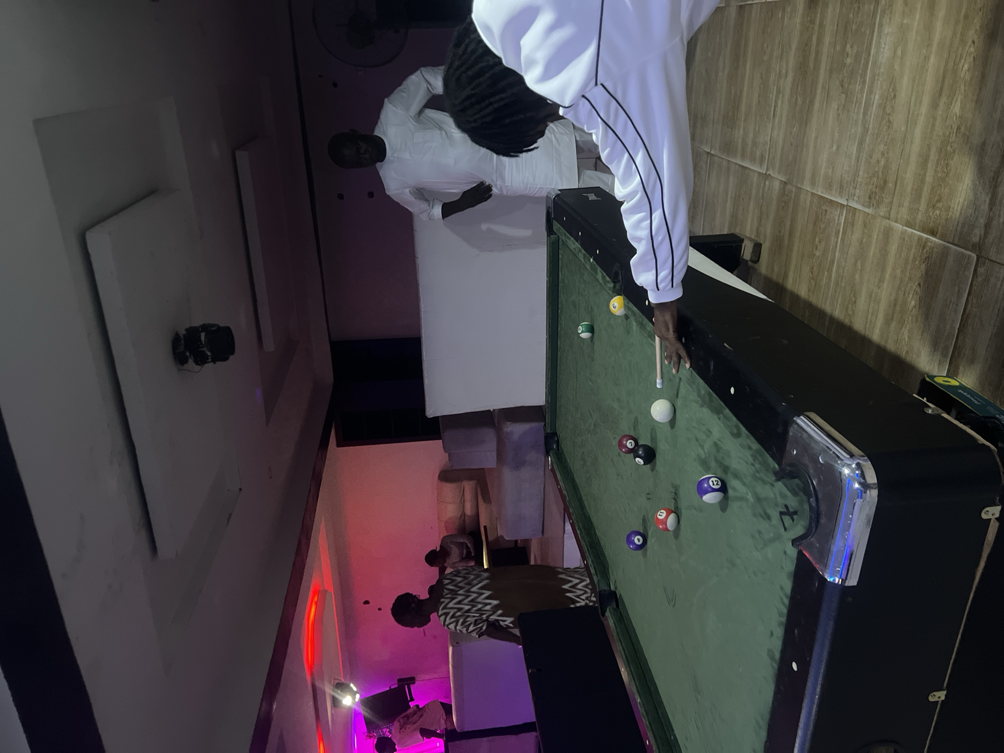 The image shows an indoor scene in Howayi, Nigeria. Two men are playing pool on a billiards table. One man is in a white outfit, and the other is in a patterned shirt. There are several billiard balls on the table. The room is illuminated with a mix of ambient and decorative lighting, including red neon lights. Other people are present in the background. The setting appears to be a leisure or entertainment space.