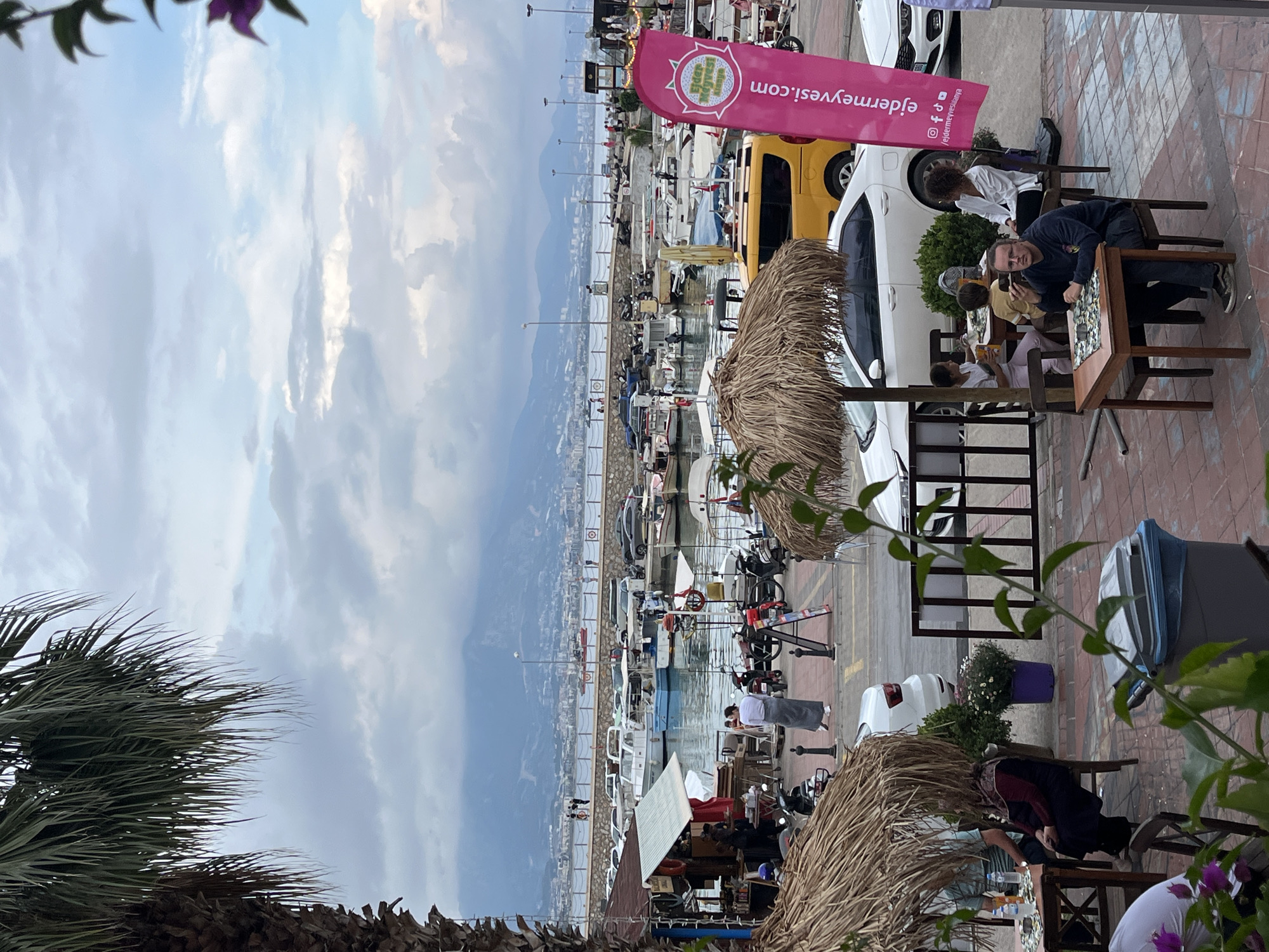 The image captures a scene in Alanya, Turkey, along the harbor. The photo appears to be taken from a slightly elevated position. A harbor with many boats is visible in the middle-ground. A mountain range runs in the background. The sky is cloudy.
In the foreground, there is outdoor seating for a restaurant or cafe. People are seated at tables, eating and engaging in conversation. There are umbrellas made of straw providing shade. Palm trees are visible in the lower left corner.
Cars are parked along the street, and people are walking nearby. A sign with Turkish writing is planted near the seating area.