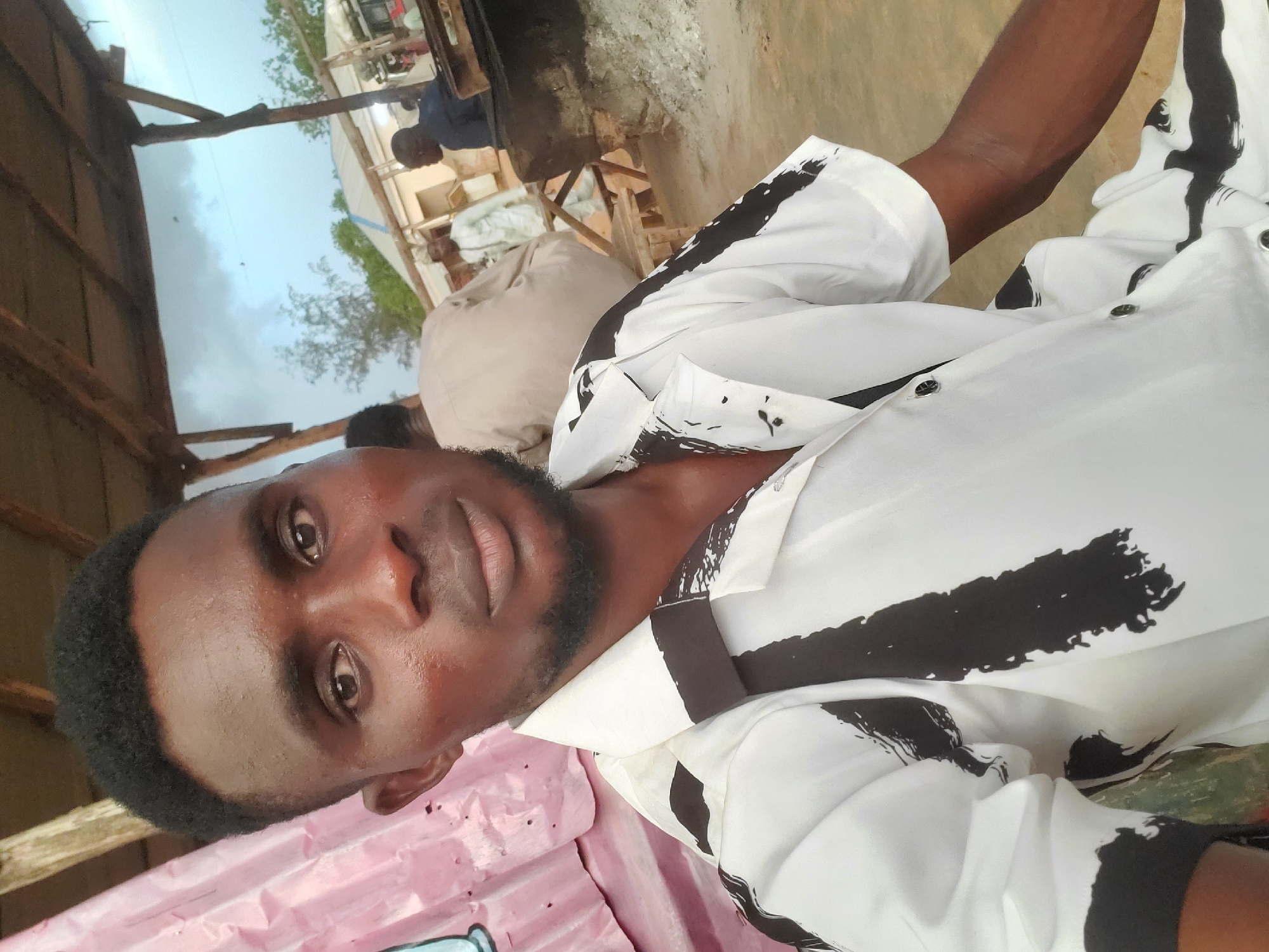 A medium shot shows a young Black man with short, dark hair and a light beard, wearing a white short-sleeved shirt with a black abstract pattern. He is looking at the camera. He is in an outdoor setting that appears to be a marketplace or roadside area in Funtua, Nigeria. In the background, other people are visible, some sitting and some standing, and there are structures that suggest stalls or shops. The sky is cloudy. The man is the primary subject.