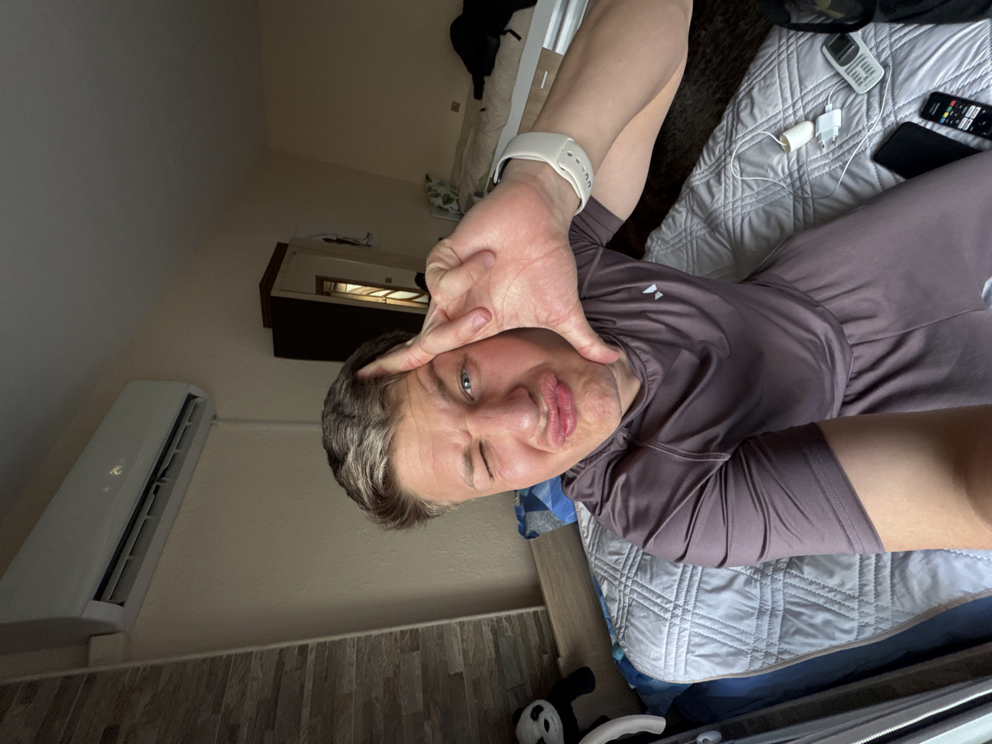 A man with short blonde hair and wearing a white watch is angled slightly to the right, making a funny face with his hand near his forehead. He is wearing a dark purple athletic shirt and shorts. The room is dimly lit. To the left is an air conditioning unit on the wall. The background includes a bed covered with a patterned gray blanket, along with various items like remotes and a phone charger. The location is likely indoors, potentially a hotel room, but the specific city of Burgas, Bulgaria cannot be confirmed with certainty.