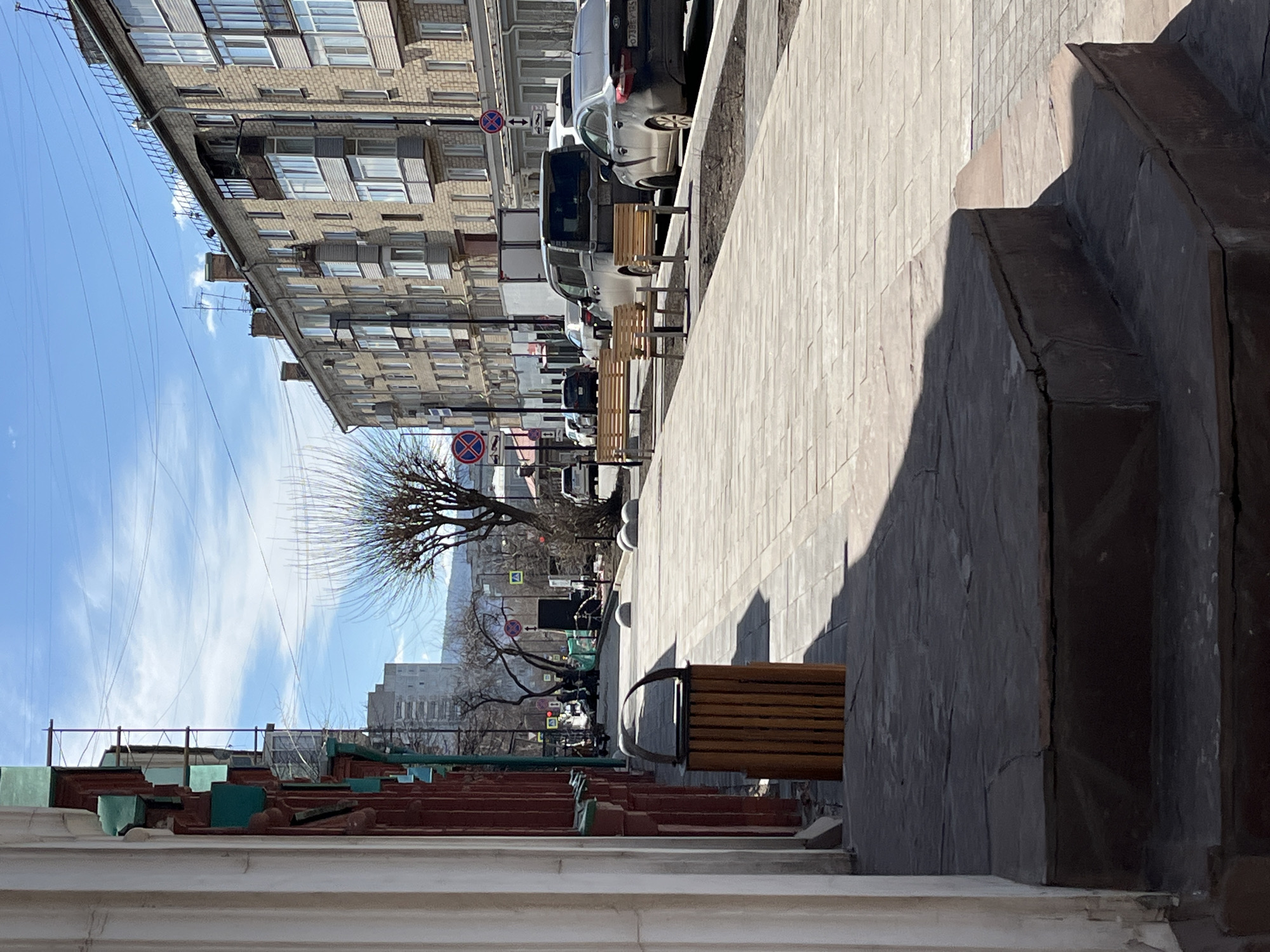 An urban street scene unfolds under a bright, sunny sky, suggesting mid-day. A multi-story brick building with numerous windows lines the left side, exhibiting architecture common in Eastern European cities. The street below it is populated by several parked cars, including a light-colored SUV and a dark sedan. A Russian license plate, "O 728 MC 93", is visible on one vehicle, providing a strong geographical clue.
A wide, paved sidewalk stretches across the central portion of the image, featuring several wooden benches with dark metal frames. Bare trees along the sidewalk indicate late winter or early spring. Various road signs are present, including a "No Stopping" or "No Parking" sign (red circle with a blue diagonal line), a blue directional arrow, and a yellow warning sign. Overhead power lines crisscross the blue sky, which has scattered wispy clouds. Antennas are visible on the rooftops of the brick building.
In the right foreground, large, dark, weathered stone or concrete structures with stepped forms cast prominent shadows. Below these, decorative elements resembling a roofline with white columns and red and green accents are visible, suggesting the facade of a building or a complex architectural feature. No people are present, lending a quiet atmosphere to the urban environment.