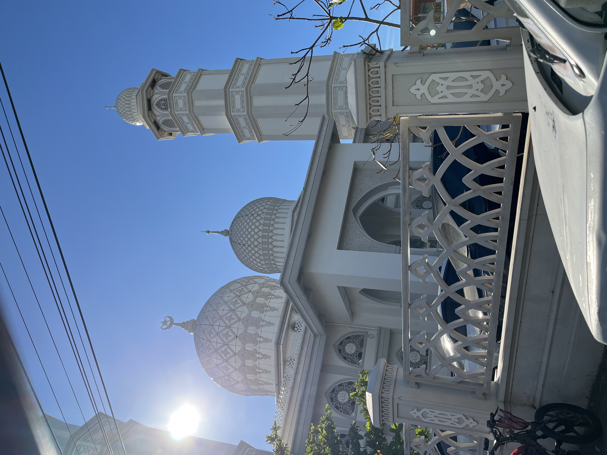 The image captures a mosque with multiple domes and a tall, slender minaret against a clear blue sky. The architecture is predominantly white, featuring intricate geometric patterns on the domes and decorative latticework on the walls and railings. A crescent moon and star symbol adorns the top of one of the smaller domes. In the foreground, a portion of a white vehicle is visible on the right, and power lines are present on the left, suggesting an urban or suburban setting. A bicycle with a red bag is parked near the fence on the right side. The sun is shining brightly, casting lens flare and creating strong highlights on the white structures, indicating it is likely daytime. The scene is serene and appears to be a place of worship in Lambaro, Indonesia. There is no visible text in the image.