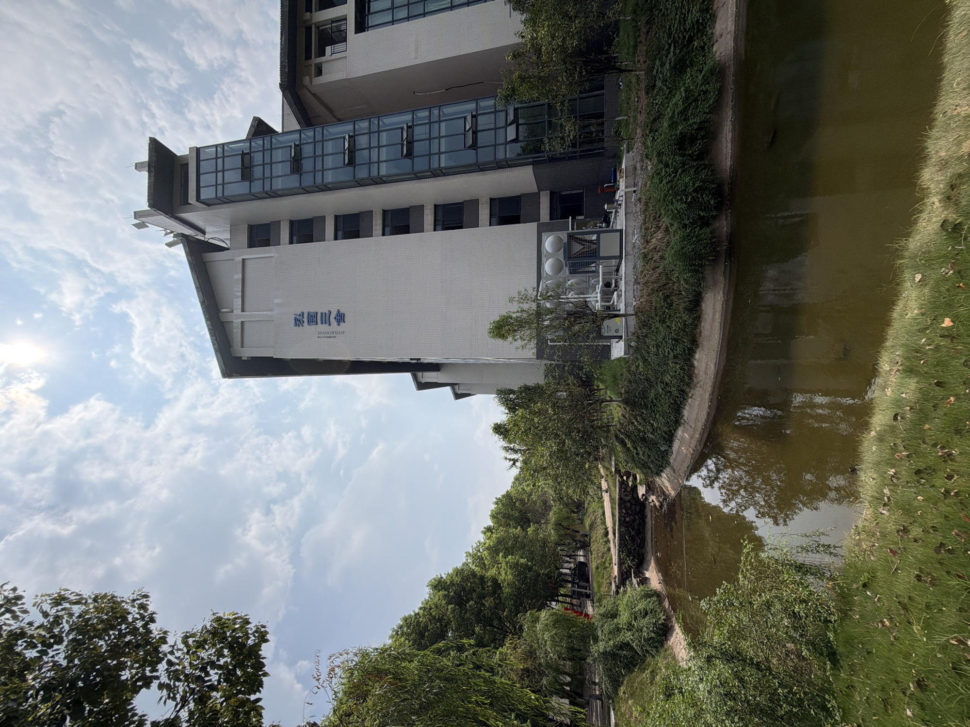 In the city of Jiangjin, China, a modern multi-story building with a glass elevator shaft stands adjacent to a narrow, man-made canal. The building's facade is white, with some sections appearing to be made of textured tiles. Blue Chinese characters are visible on the upper part of the building. A cluster of industrial equipment, including what appears to be air conditioning units and a water tank, is situated at the base of the building. The canal is surrounded by lush greenery, with trees and bushes lining its banks. A small stone bridge crosses the canal in the mid-ground. The water in the canal is murky and reflects the surrounding foliage and the sky. The sky is partly cloudy, with bright sunlight filtering through. The image appears to have been taken during the daytime. There are no people visible in the scene.