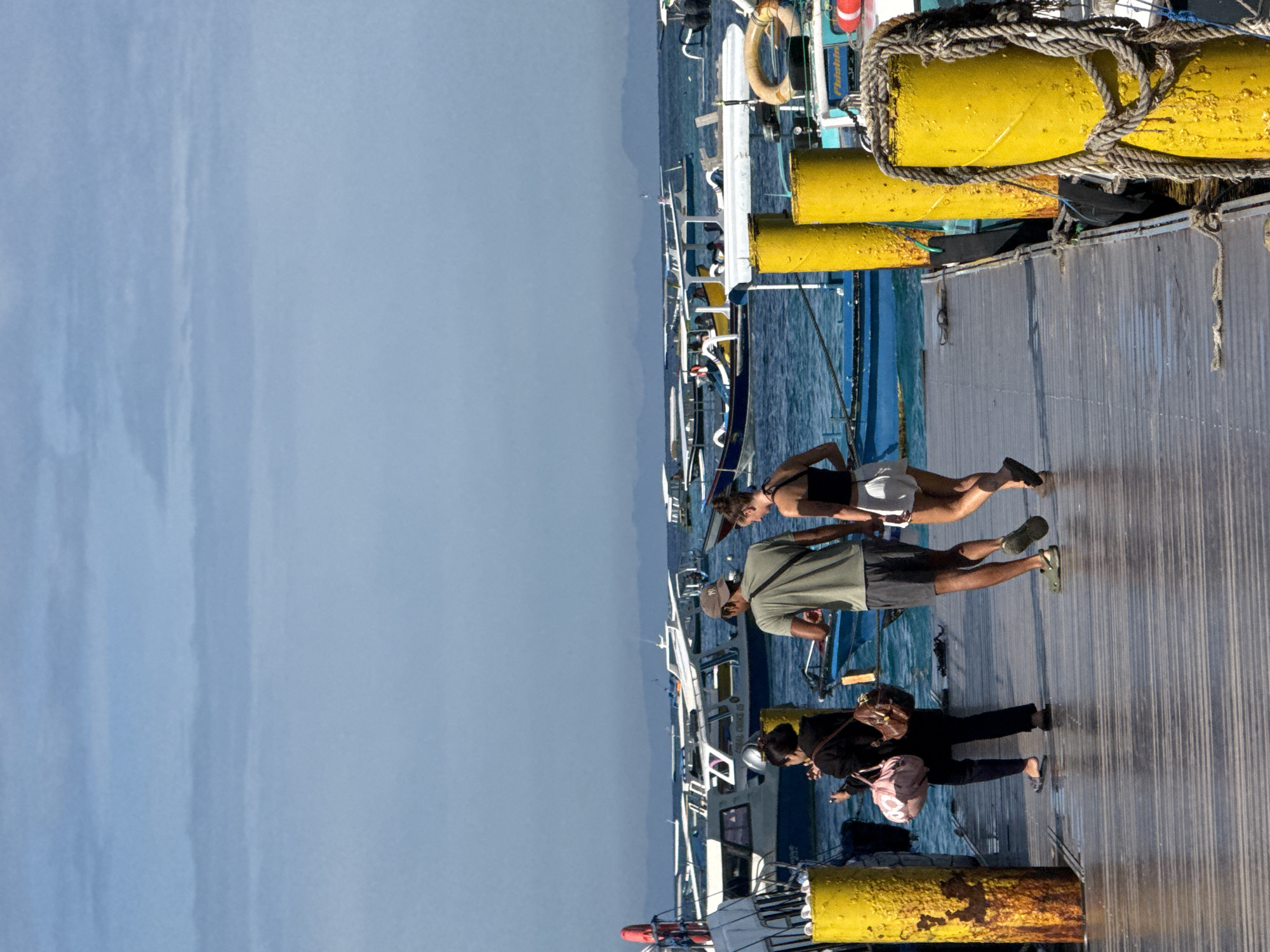 The image captures a vibrant daytime scene at a wooden pier, bustling with people and boats. The perspective is from the pier, looking down its length towards a hazy horizon.
On the right side of the image, heavy yellow cylindrical buoys, weathered and rusty in places, are secured along the edge of the dock with thick ropes. Adjacent to the pier, numerous boats are moored in the blue water. These are predominantly white passenger boats, many featuring blue accents and often equipped with outriggers, characteristic of vessels used for tours or transport in coastal regions. On one boat, a white and orange lifebuoy is visible, and faint text, including "OCEAN CRUISE", can be discerned.
Several individuals are present on the dock. Towards the foreground and right, a person in dark clothing carries a light pink backpack, standing beside another person whose head is partially visible near a boat. Further down the pier, a man in a green t-shirt, dark shorts, a baseball cap, and green sandals walks away from the camera, alongside a woman wearing a dark top and light shorts.
The water appears calm, reflecting the clear sky above. In the distant background, a low, hazy landmass (likely an island or coastline) is faintly visible under a light blue sky. The overall scene suggests a busy port or tourist hub, possibly indicating preparations for or return from boat excursions. The bright lighting suggests it's a sunny day.