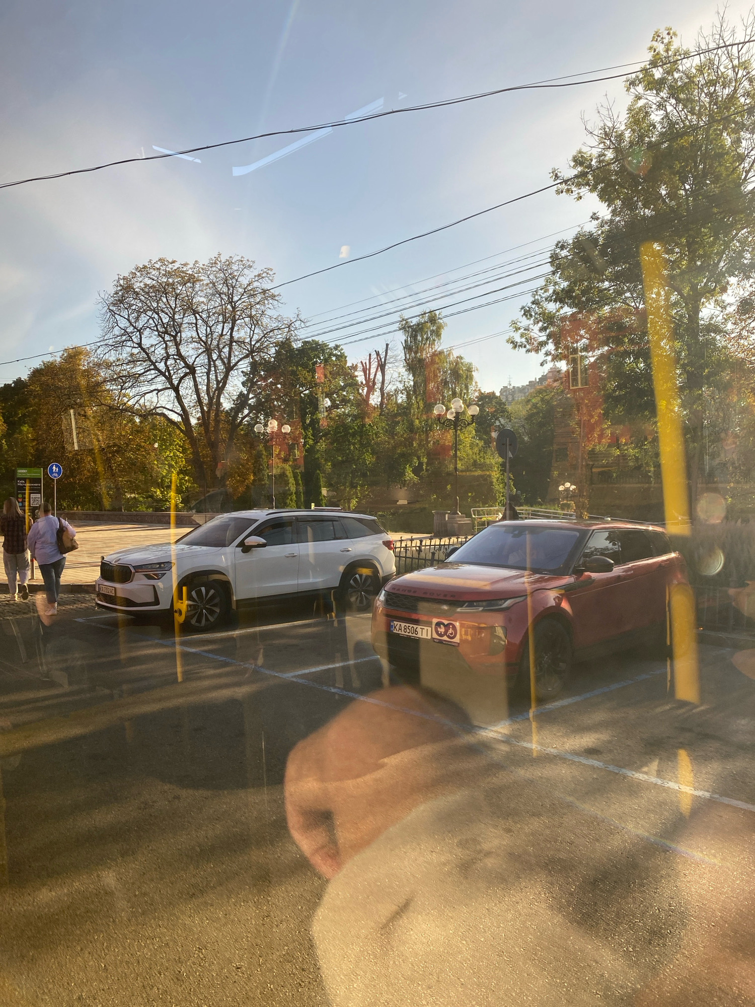 In an outdoor urban setting under a bright, clear sky, two cars are parked in designated spots. The car on the left is a white SUV, while the car on the right is a red Range Rover. Two individuals are walking away from the camera near the white SUV. One person is wearing a plaid shirt and jeans, and the other is wearing a light-colored top and pants. Trees with some autumn foliage are visible in the background, along with streetlights and power lines. The scene appears to be captured through a window, as indicated by reflections on the surface.