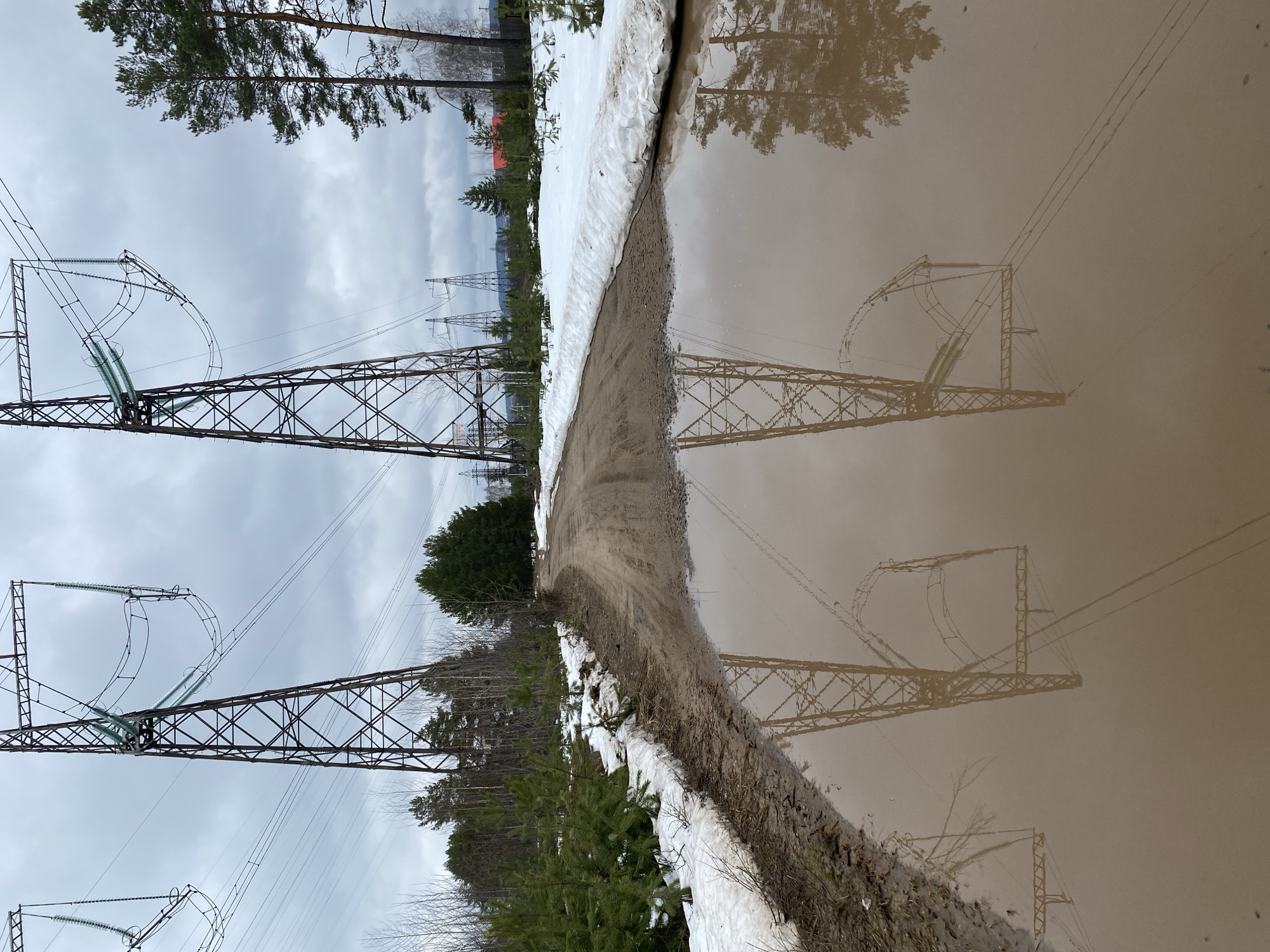The image depicts a rural scene near Noviy, Russia, with power lines dominating the landscape. The ground is partially covered in snow, with a dirt path running along a large puddle reflecting the power line towers and surrounding trees. The sky is overcast.