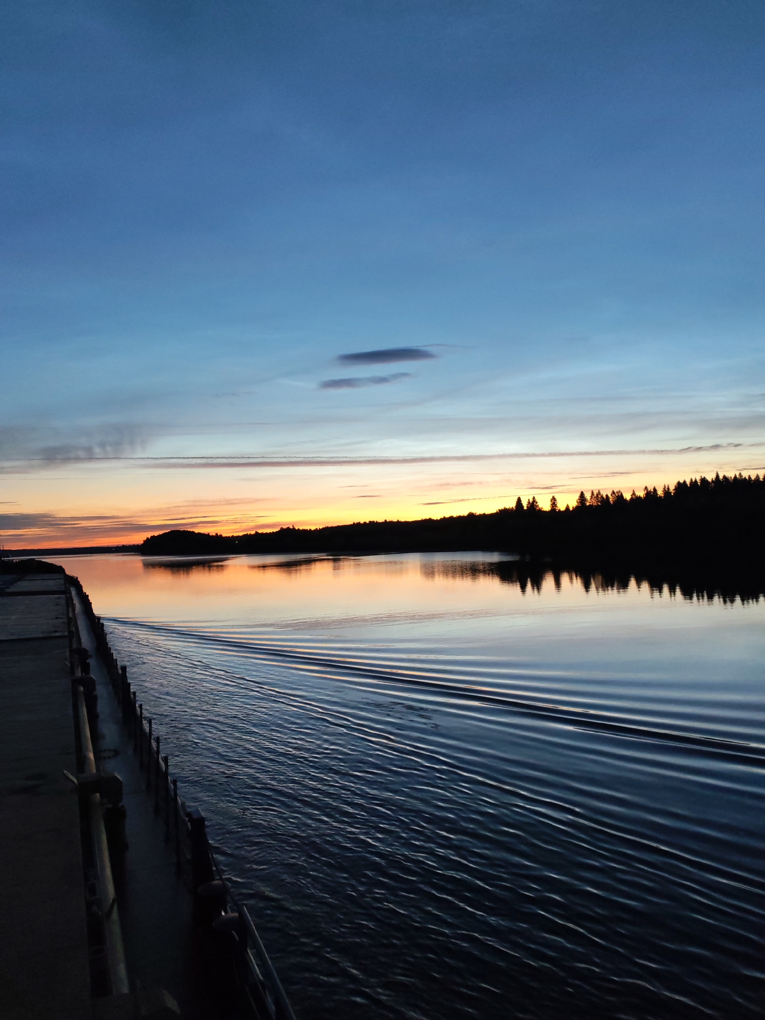 The image depicts a serene sunset over a body of water, likely a lake or a wide river, in Matveevskoe, Russia. The sky transitions from a deep blue at the top to hues of orange and yellow near the horizon, with subtle clouds streaking across. Silhouetted against the vibrant sky is a treeline of dense coniferous forest, forming a dark, jagged edge along the distant shore. In the foreground, a concrete pier or dock extends into the water from the left side of the frame. The water itself is calm, with the exception of ripples spreading outwards from the pier, suggesting movement or a boat passing by. The reflection of the sky and the treeline is visible on the water's surface, creating a symmetrical effect. The overall mood is tranquil and picturesque, capturing the quiet beauty of a rural sunset. There are no people or animals visible in the scene.