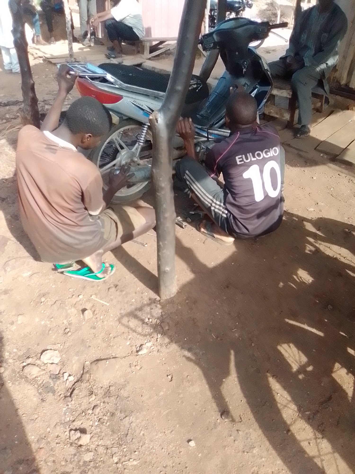 Two young men are working on a motorcycle in Pantame, Nigeria. The man on the left, wearing a brown t-shirt and shorts, is focused on the rear wheel of the motorcycle. He is wearing green flip-flops. The man on the right, wearing a dark t-shirt with "EULOGIO 10" printed on the back and striped track pants, is also attending to the motorcycle, positioned closer to its front. He is wearing sandals. The motorcycle is silver and black. The ground is dirt and gravel, with some rocks and debris scattered around. In the background, other people and parts of structures can be seen, suggesting an outdoor workshop or open-air setting. The lighting indicates it is daytime, with shadows cast on the ground.