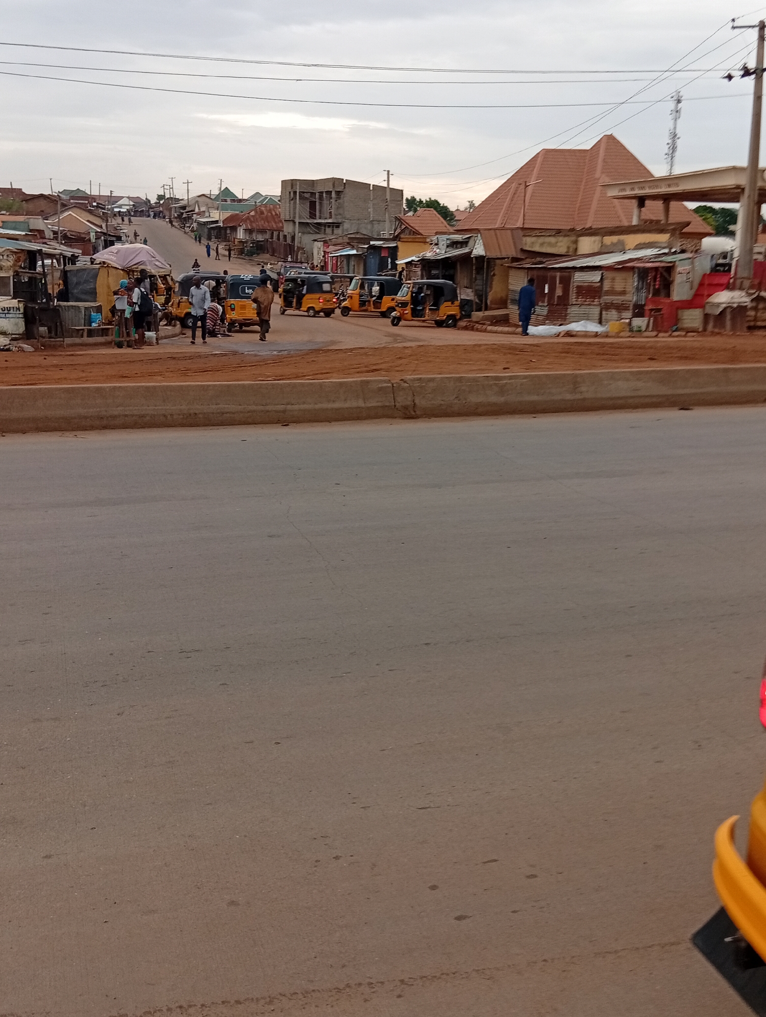 The image depicts a street scene in Nassarawa, Nigeria. The scene is dominated by a wide, asphalt road in the foreground. A concrete barrier separates the road from a bustling roadside area. Numerous yellow auto-rickshaws (tuk-tuks) are visible along the roadside, along with several people standing and interacting in small groups. Makeshift stalls and shops line the street behind the auto-rickshaws. A building under construction is also visible. Overhead power lines are present. The sky is overcast.
