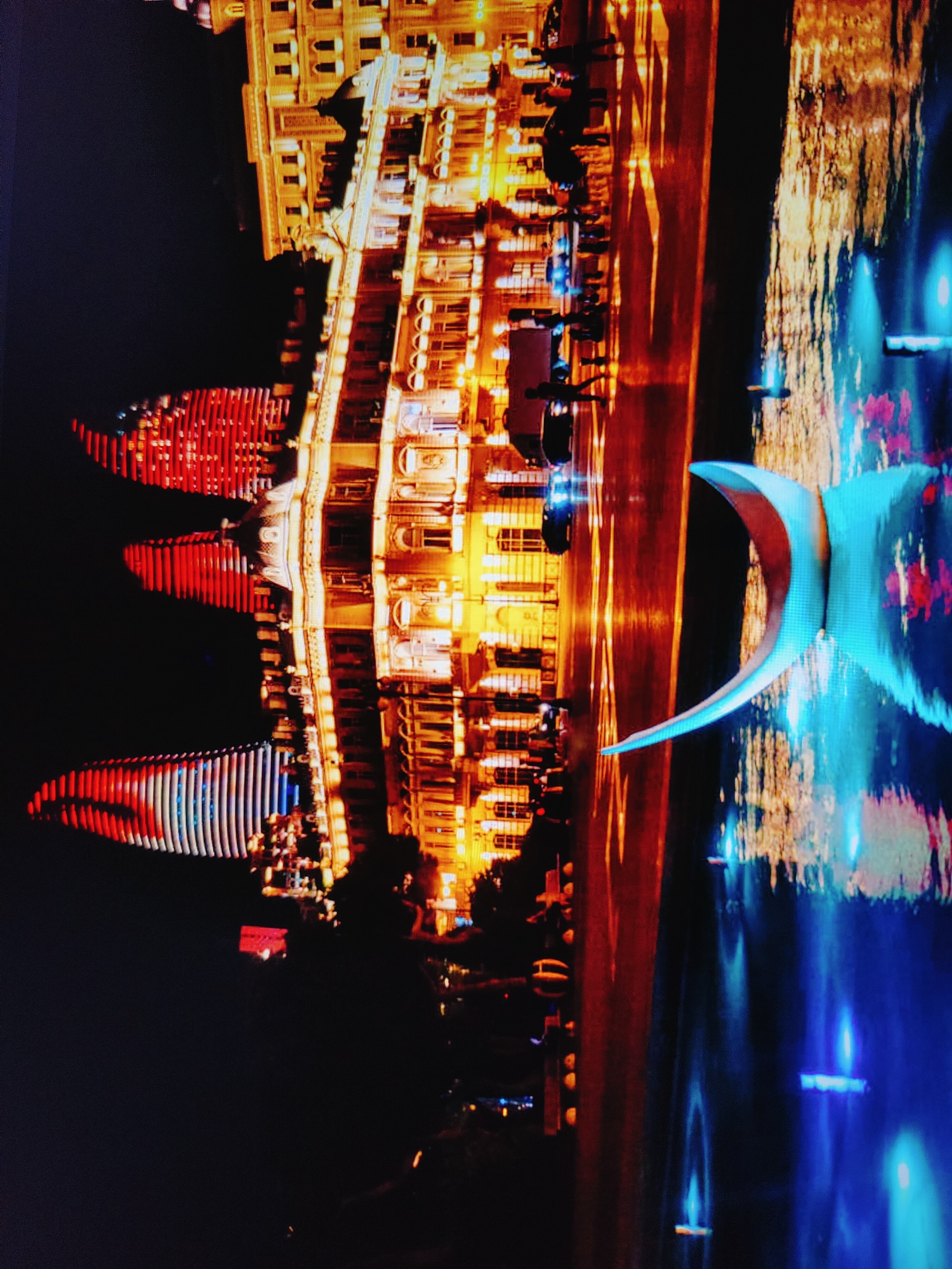 The image depicts a nighttime scene in Baku, Azerbaijan, showcasing a blend of architectural grandeur and vibrant urban illumination. A large, ornately lit building with numerous windows and balconies dominates the right side of the frame, its facade glowing with warm, yellow light. In front of this building, a body of water reflects the lights, creating a shimmering surface.
On the left side, three large, sculptural structures are illuminated with bright red and white lights, resembling flames or stylized flames. These structures appear to be part of a larger installation or display, with their dynamic lighting contrasting sharply with the static architecture. The sky is dark, indicating late evening or nighttime.
The scene also includes elements of urban life, such as the reflections of car headlights on a wet surface, suggesting a road or pathway alongside the water. Silhouettes of people are visible along the edge of the illuminated building and near the red structures, implying a public space with pedestrian activity. A distinctive blue, curved sculpture is also visible in the water, adding another artistic element to the composition. The overall impression is of a modern, well-lit cityscape with significant artistic and architectural features.