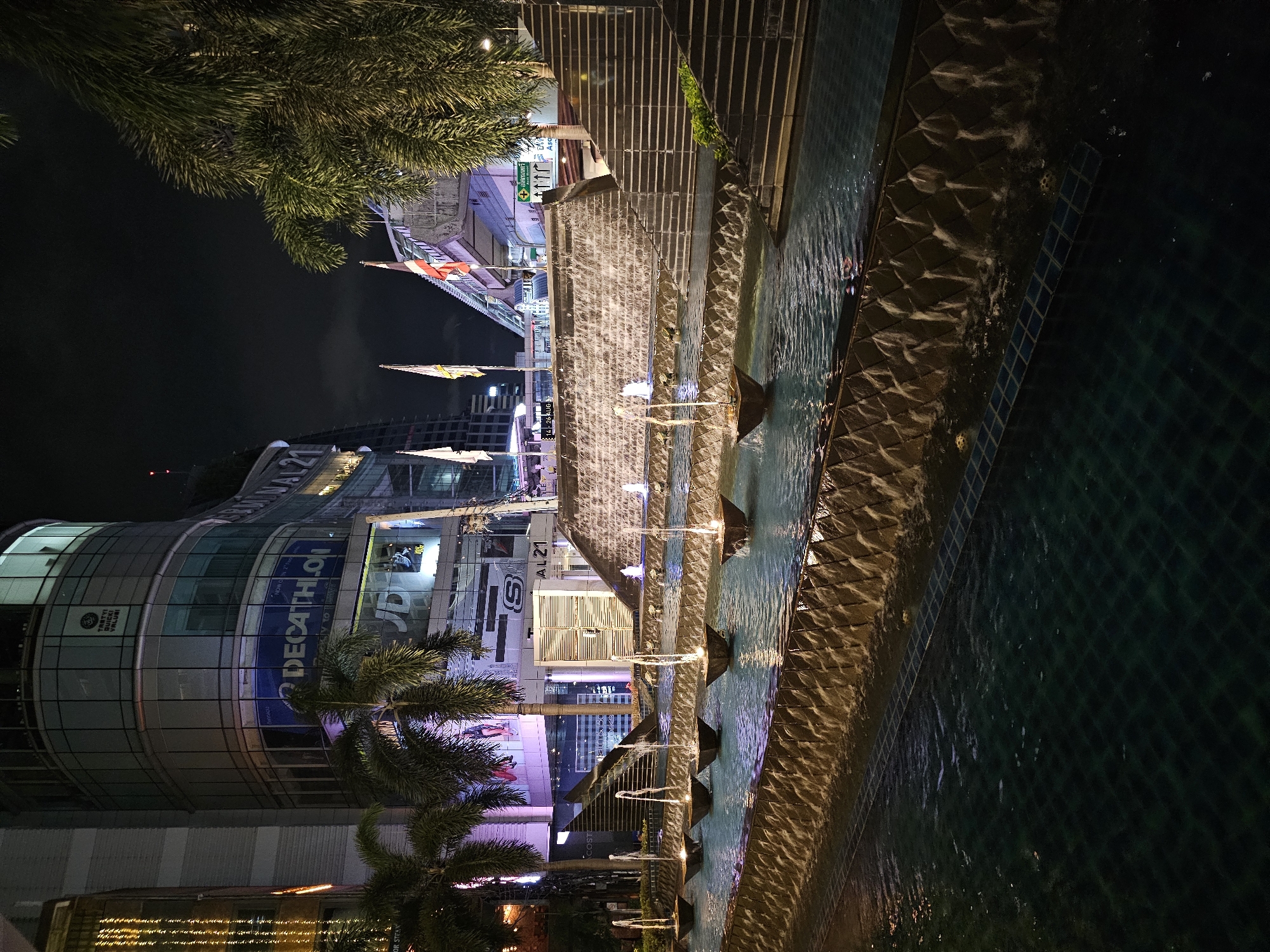 The image depicts a nighttime scene in Bangkok, Thailand, featuring a multi-tiered water feature with fountains in front of a modern building complex. The building, likely a shopping mall or commercial center, has a glass facade with illuminated signage, including "DECATHLON" and "JD SPORTS". Palm trees are visible on the left side of the frame. The water feature consists of several levels of cascading water, with fountains spraying upwards from individual bowls. Stairs are visible on the right side of the water feature, leading up towards a skywalk or elevated walkway. Flags are flying from poles on the elevated structure. The overall atmosphere is urban and illuminated by artificial lights. There are no people visible in the image.