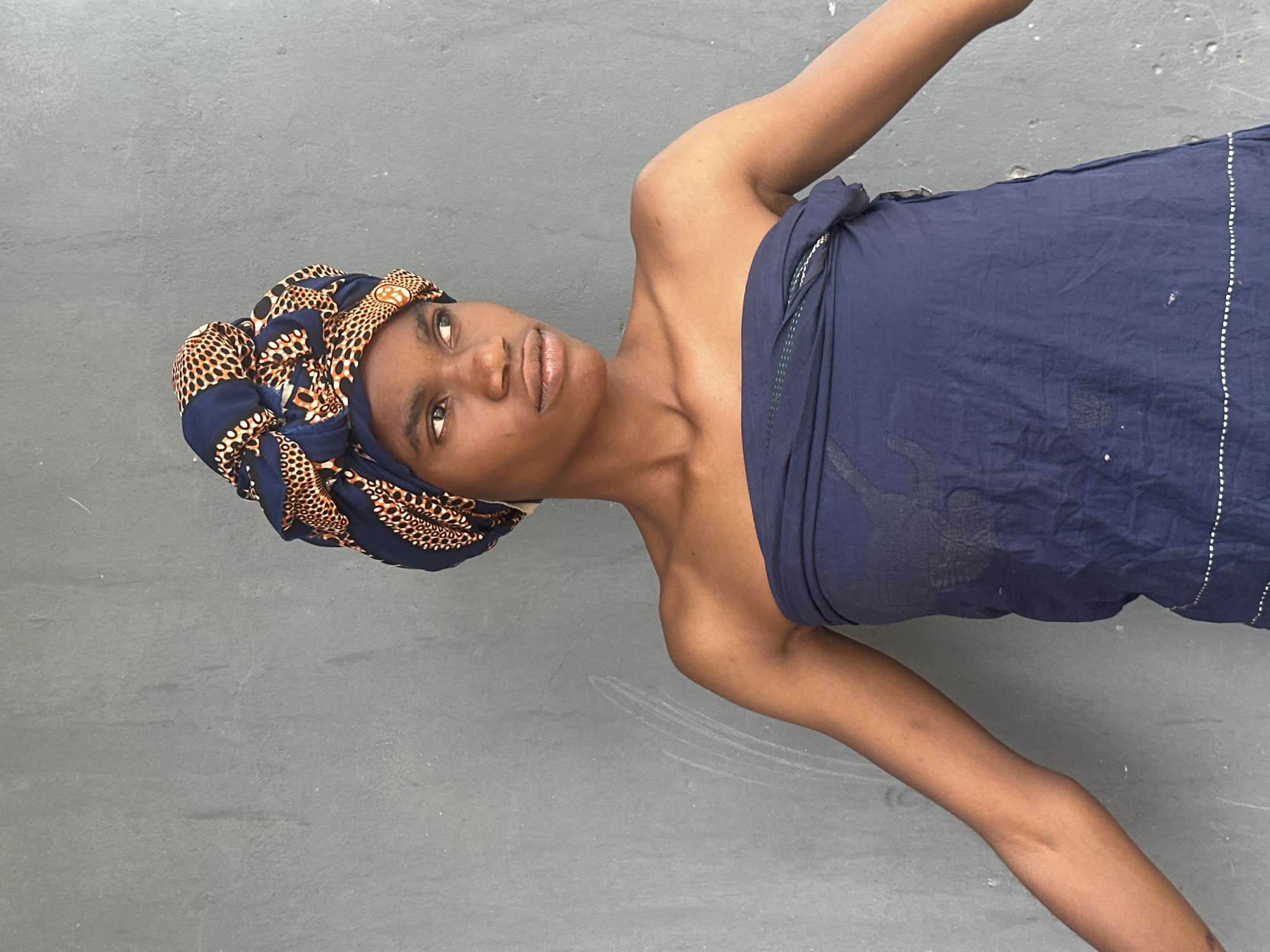 A young person with dark skin, wearing a blue and orange patterned headwrap and a dark blue strapless garment, stands against a textured grey wall. Their arms are extended outwards. The background appears to be an outdoor, possibly urban setting in Badagry, Nigeria, suggested by the grey concrete wall. The lighting is bright, casting soft shadows.