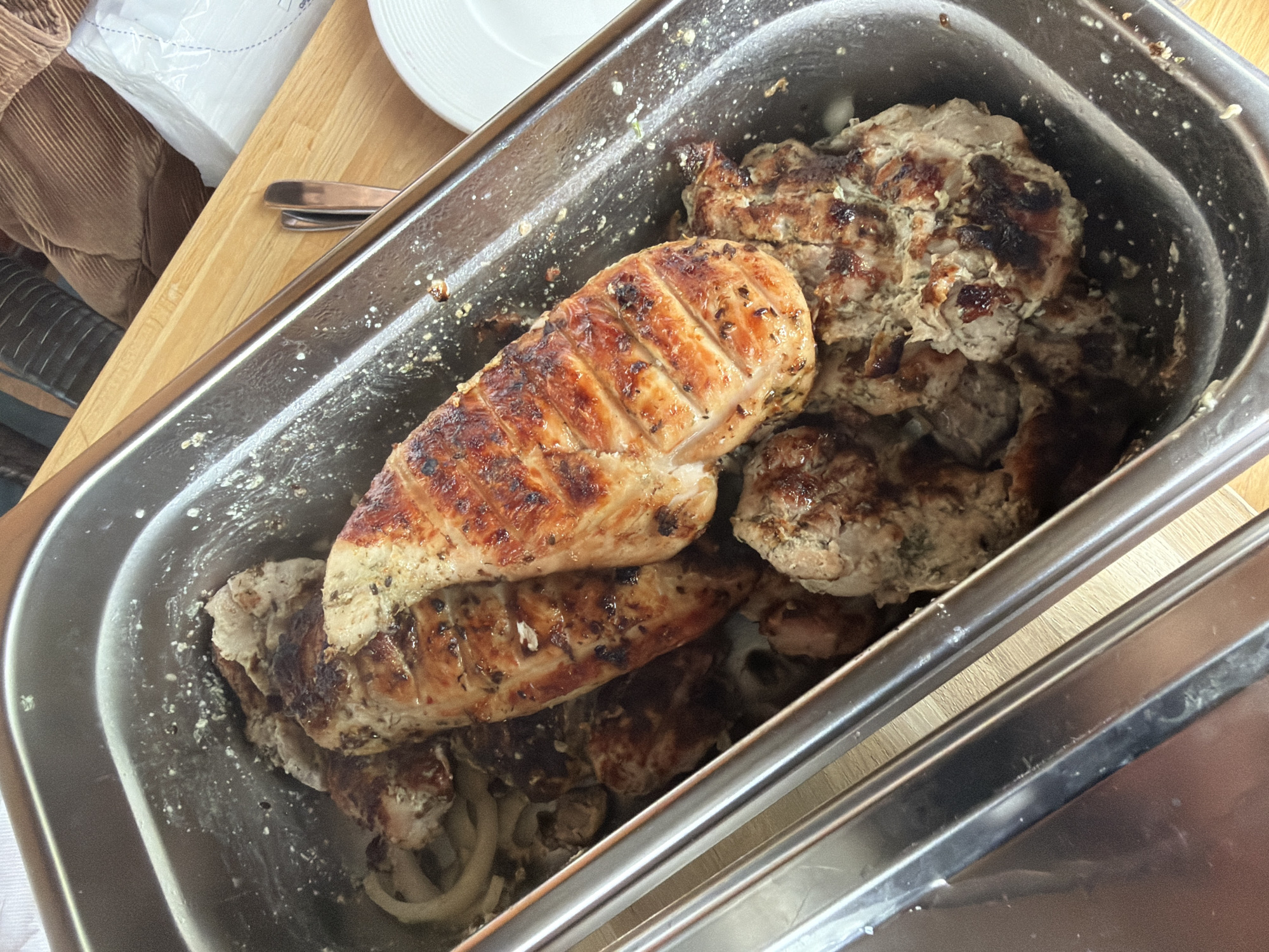 A close-up, high-angle shot shows grilled chicken in a rectangular stainless steel container. The chicken pieces are varied in size and shape, with some appearing to be breasts and others smaller, possibly thighs or cutlets. The surface of the chicken exhibits a golden-brown sear with grill marks visible on some pieces. The container also contains sliced onions and herbs mixed with the chicken. The background suggests an outdoor setting, possibly a picnic or barbecue, with a wooden surface and blurred elements of furniture and a white paper product. The scene does not provide any specific landmarks or identifiable features to pinpoint a location within Baloži, Latvia.