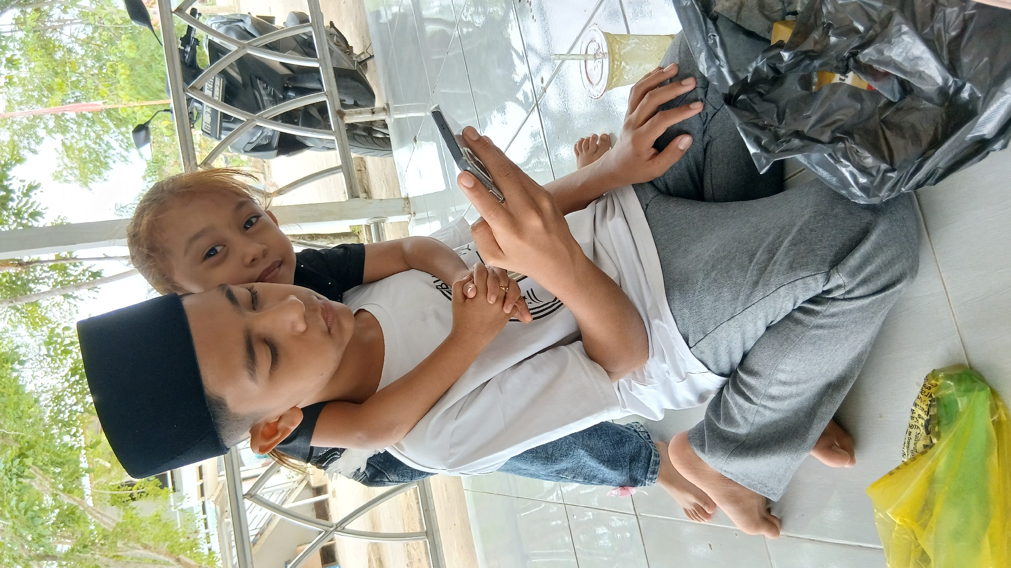 A young boy wearing a black peci and a white t-shirt is sitting on the floor, looking at his phone. He has his arms crossed, and his hands are resting on his lap. A young girl, with blonde hair and wearing a black shirt, is standing behind him and has her arms wrapped around his neck. She is leaning her head on his shoulder and looking at the camera. They appear to be in an outdoor setting, possibly a porch or patio, with a tiled floor and a metal railing in the background. There is a motorcycle parked behind the railing, and trees and foliage can be seen in the distance. A plastic bag and a cup with a straw are on the floor near the boy. The image seems to be taken in Matangben, Indonesia.