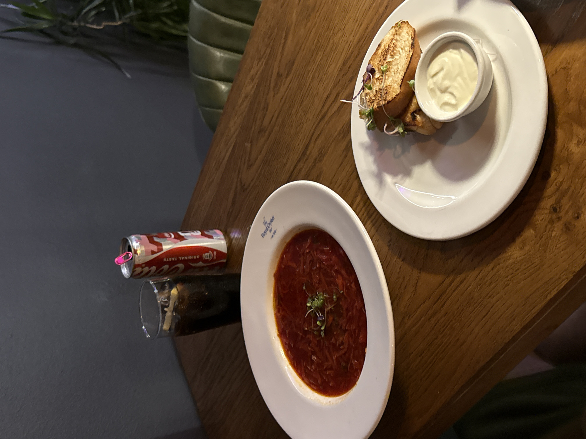 On a wooden table, an overhead shot reveals a partially consumed can of Coca-Cola next to a glass of dark liquid, possibly Coca-Cola itself, and a white bowl containing a red soup garnished with sprouts. A second white plate holds what appears to be a halved sandwich or roll accompanied by a small bowl of white sauce. The angle suggests a casual dining setting. The location, deduced from the "Royal Oyster Bar" on the soup bowl, is likely Cape Town, South Africa, given that this establishment is known to exist there. The scene depicts a meal in progress, with no individuals explicitly visible.