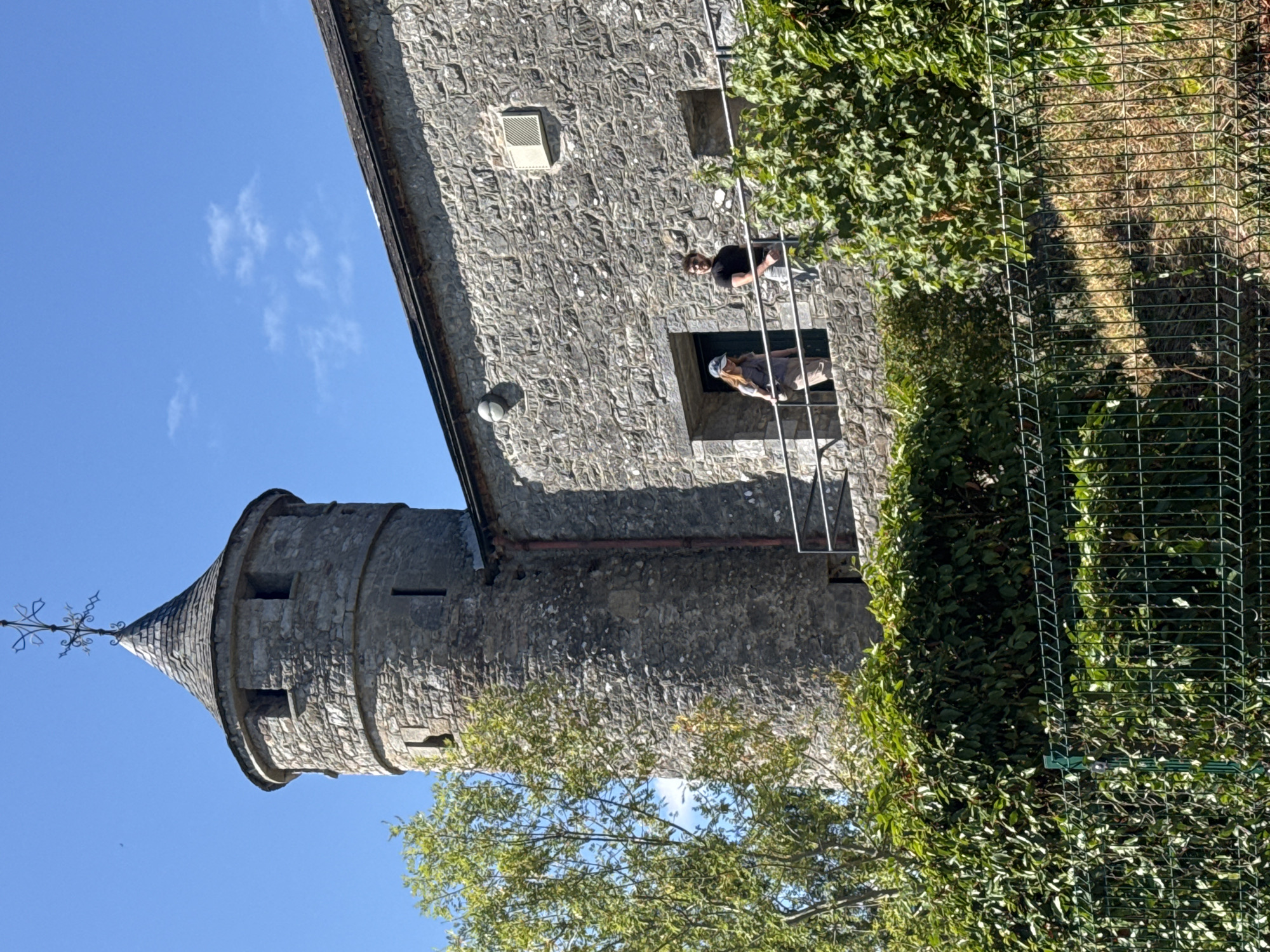 A stone tower with a conical roof stands tall against a clear blue sky. Two people, a woman wearing a light-colored top and shorts, and a man in a black t-shirt, are on a balcony overlooking a lush green landscape. The woman is on the left, and the man is on the right. A metallic railing runs along the balcony. The tower is part of a larger stone building, possibly a historical structure. In the foreground, a green metal fence encloses dense green foliage. The scene is bathed in sunlight, casting shadows. This is a view of Cahir Castle in Cahir, Ireland.