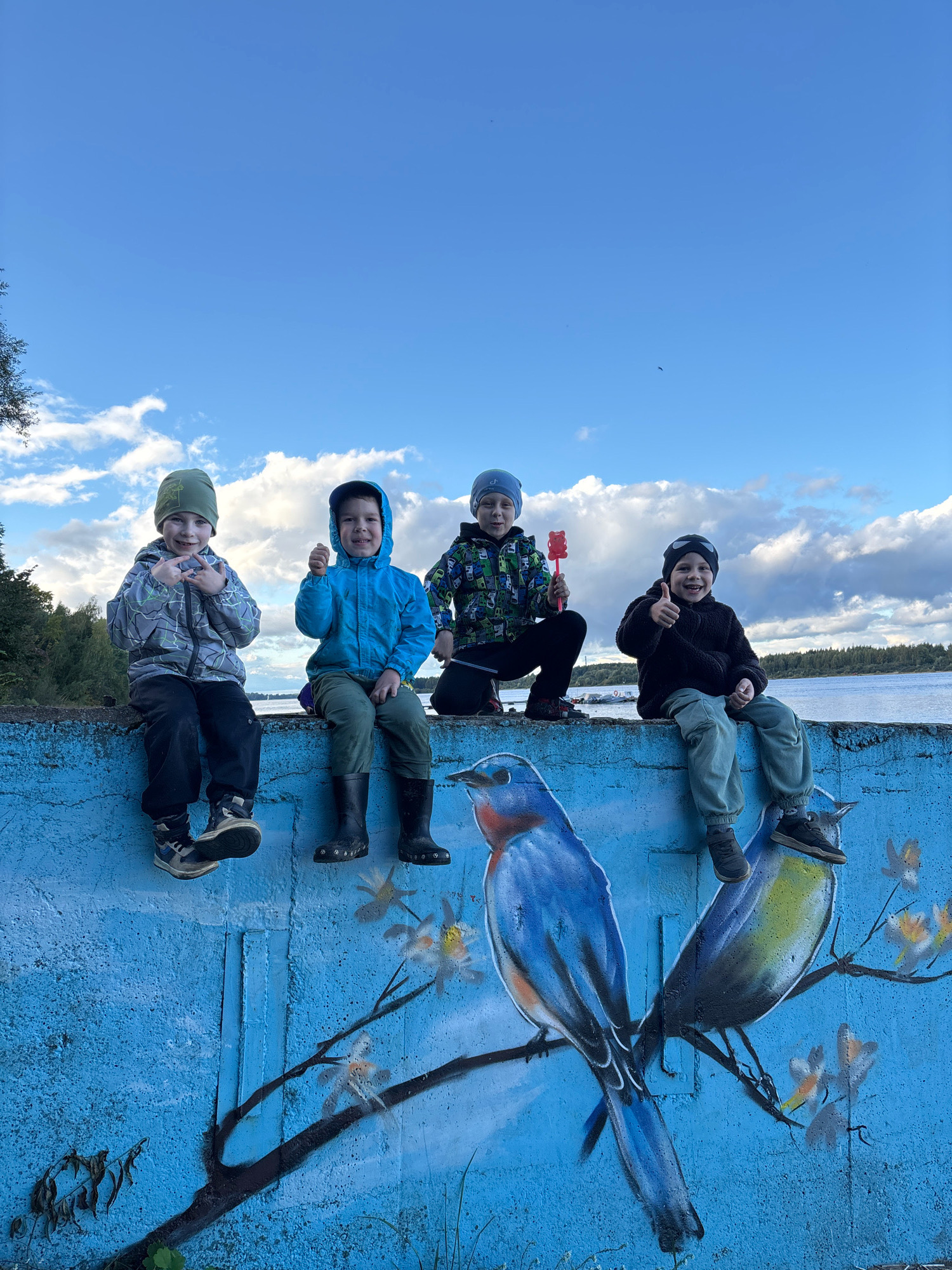 Four young boys are sitting on a blue concrete wall with a mural of birds. The boy on the far left is wearing a grey and black patterned jacket, black pants, and a green beanie. He is making a peace sign with his fingers. The second boy from the left is wearing a blue hooded jacket, green pants, and black boots. He is giving a thumbs-up. The third boy from the left is wearing a green, black, and white patterned jacket, black pants, and a blue beanie. He is holding a red toy. The boy on the far right is wearing a black fuzzy jacket, light green pants, and a black beanie. He is also giving a thumbs-up. The wall is painted with a mural of two birds on a branch, one blue and one yellow and gray. In the background, there is a body of water, trees, and a cloudy sky. The scene appears to be outdoors in a park or recreational area. The location is identified as Zhukovo, Russia.