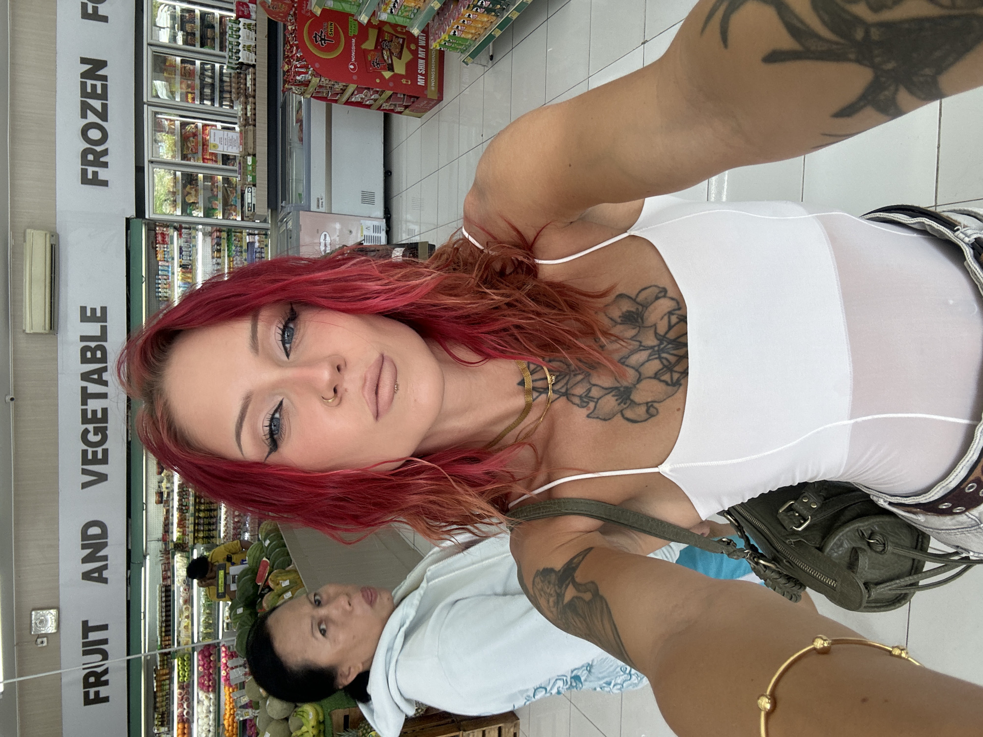 Here is a description of the image:
The image shows a woman with pink hair wearing a white top and jeans with a studded belt, taking a selfie in what appears to be a grocery store or supermarket in Kuta Selatan, Indonesia. She has a large tattoo on her chest and another on her arm, as well as several piercings on her face. Behind her, another woman, possibly her mother, is partially visible, looking up towards the camera.
The background features shelves stocked with various grocery items, refrigerators filled with frozen foods, and signage written in English, including the words "FRUIT AND VEGETABLE" and "FROZEN". A display of Shin Ramyun instant noodles can also be seen. The general environment suggests a typical shopping experience in a moderately sized store.