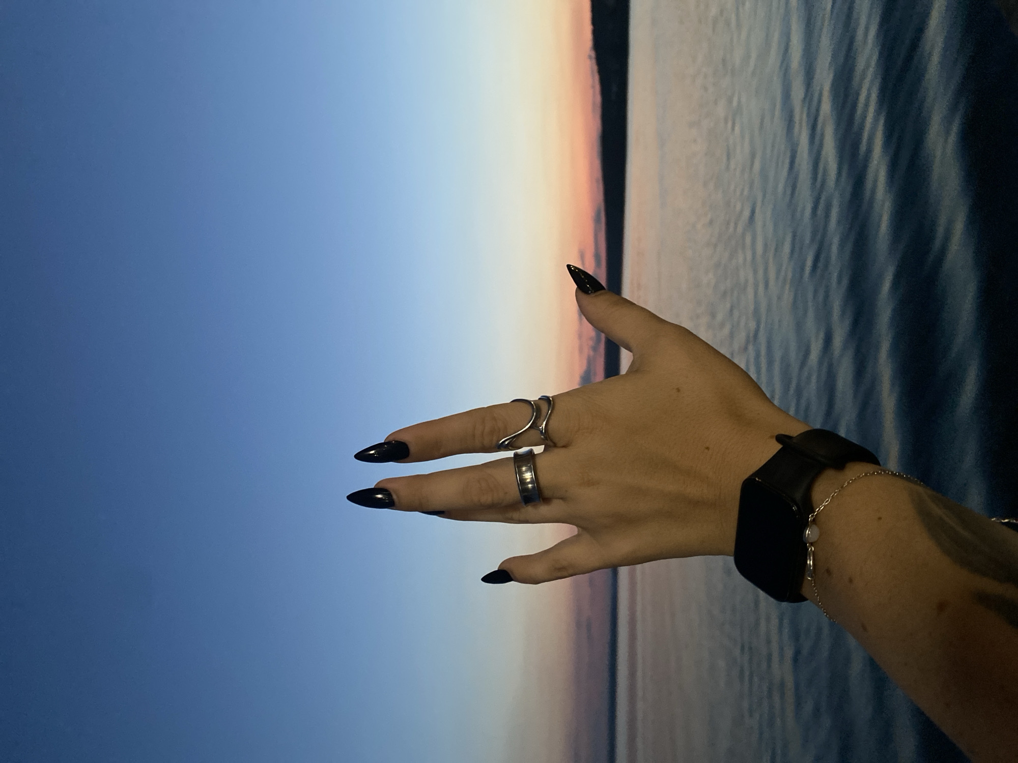 A close-up, Dutch angle shot captures a person's left hand, adorned with black, pointed fingernails and several rings, raised against a dusky sky and calm body of water. The hand is positioned in the foreground, with the wrist bearing a black smartwatch and a delicate bracelet. The background features a gradient sky transitioning from deep blue at the top to hues of pink and orange near the horizon, where a dark, silhouetted landmass is visible. The water exhibits subtle ripples, reflecting the ambient light. The image is set at dusk, likely over the Votkinsk Reservoir in Votkinsk, Russia, given the specific geographical reference.