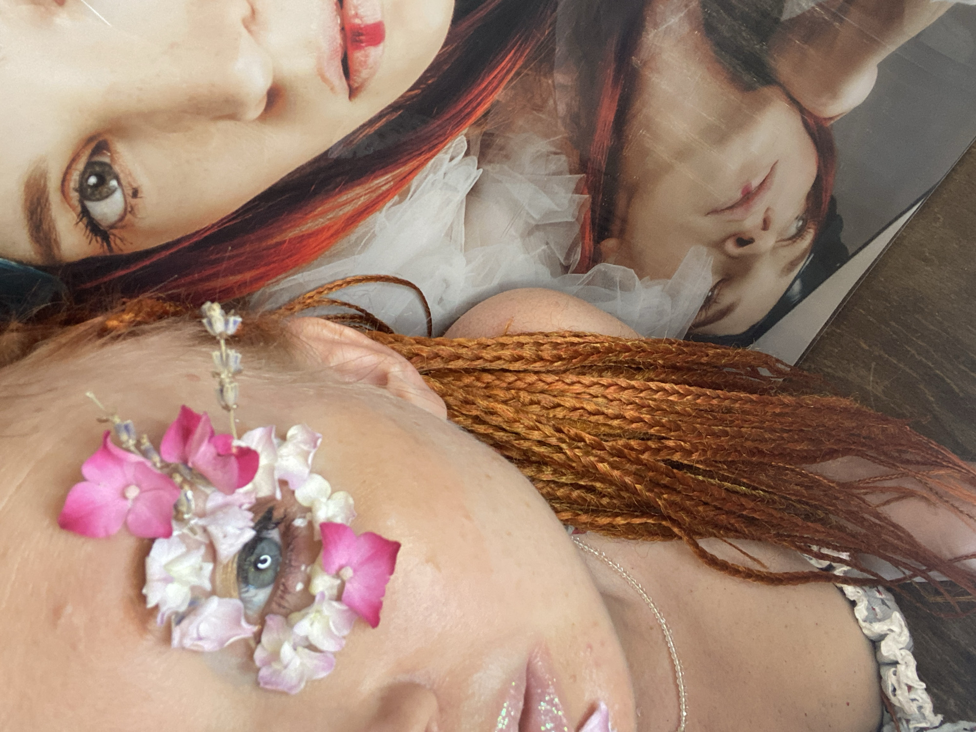 The image shows a close-up of a person's face with flowers and petals arranged around their eye. The person has auburn braids and is wearing glittery lip gloss. In the background, there is a printed image of another person's face with red streaks in their hair and red lipstick. The scene appears to be indoors, possibly a studio or a creative space. There is no visible indication of the city of Sochi, Russia.