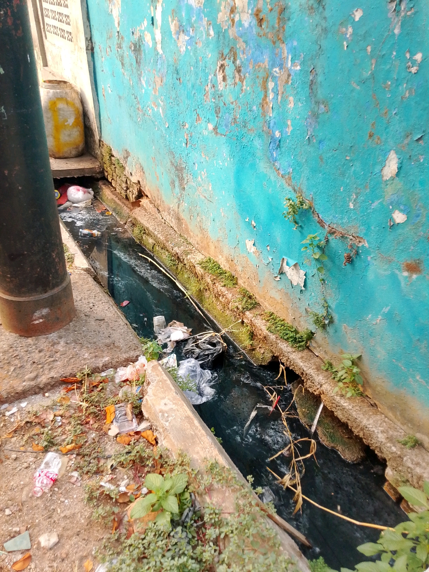 This image depicts a clogged drainage canal in East Jakarta, Indonesia, laden with refuse. The canal, a dark, stagnant channel, is choked with plastic bags, bottles, and other debris. The surrounding environment is unkempt, with a peeling turquoise wall on one side and a concrete pathway littered with trash on the other. A large black pipe stands prominently on the left, and a yellow barrel is visible near the building's foundation. The scene suggests a lack of waste management and sanitation in the area.