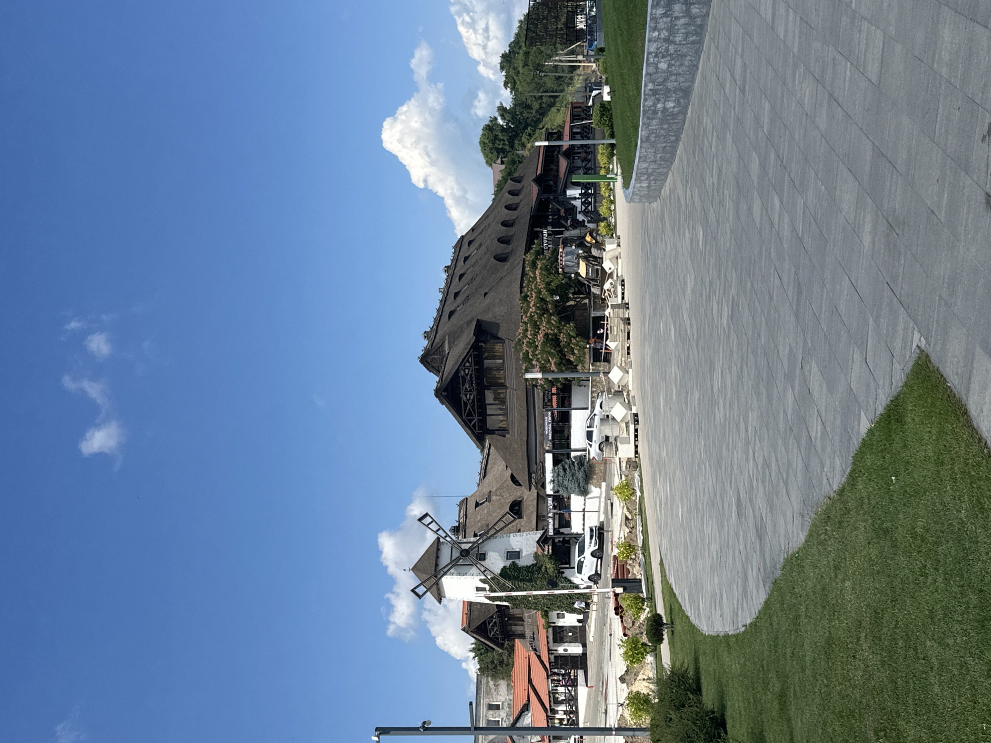 In Taman, Russia, a sun-drenched outdoor scene unfolds with a focus on a complex of buildings featuring traditional thatched roofs and a prominent windmill. The architecture suggests a restaurant or tourist establishment. In the foreground, a neatly manicured lawn curves alongside a paved walkway made of gray rectangular stones, bordered by a low, gray stone wall. Beyond the walkway, the buildings are visible, with the white windmill standing out against the blue sky dotted with fluffy white clouds. Construction materials, including pallets and cylindrical pipes, are present near the buildings, indicating ongoing work. Several cars are parked in the vicinity, including a white SUV and a silver sedan. One person is walking near the windmill, and a few individuals are visible near construction equipment, including a yellow tractor, on the right side of the frame. Trees and shrubbery are interspersed throughout the scene, adding greenery to the environment.